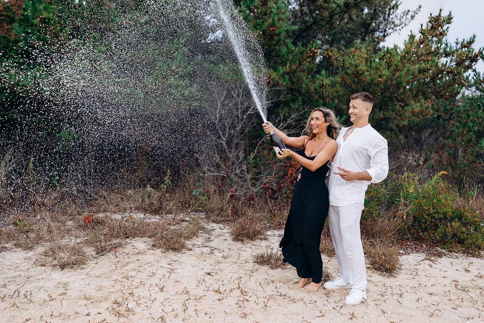 Couple on sandy beach surrounded by greenery, woman in black dress popping champagne bottle with spray, man in white outfit holding glass