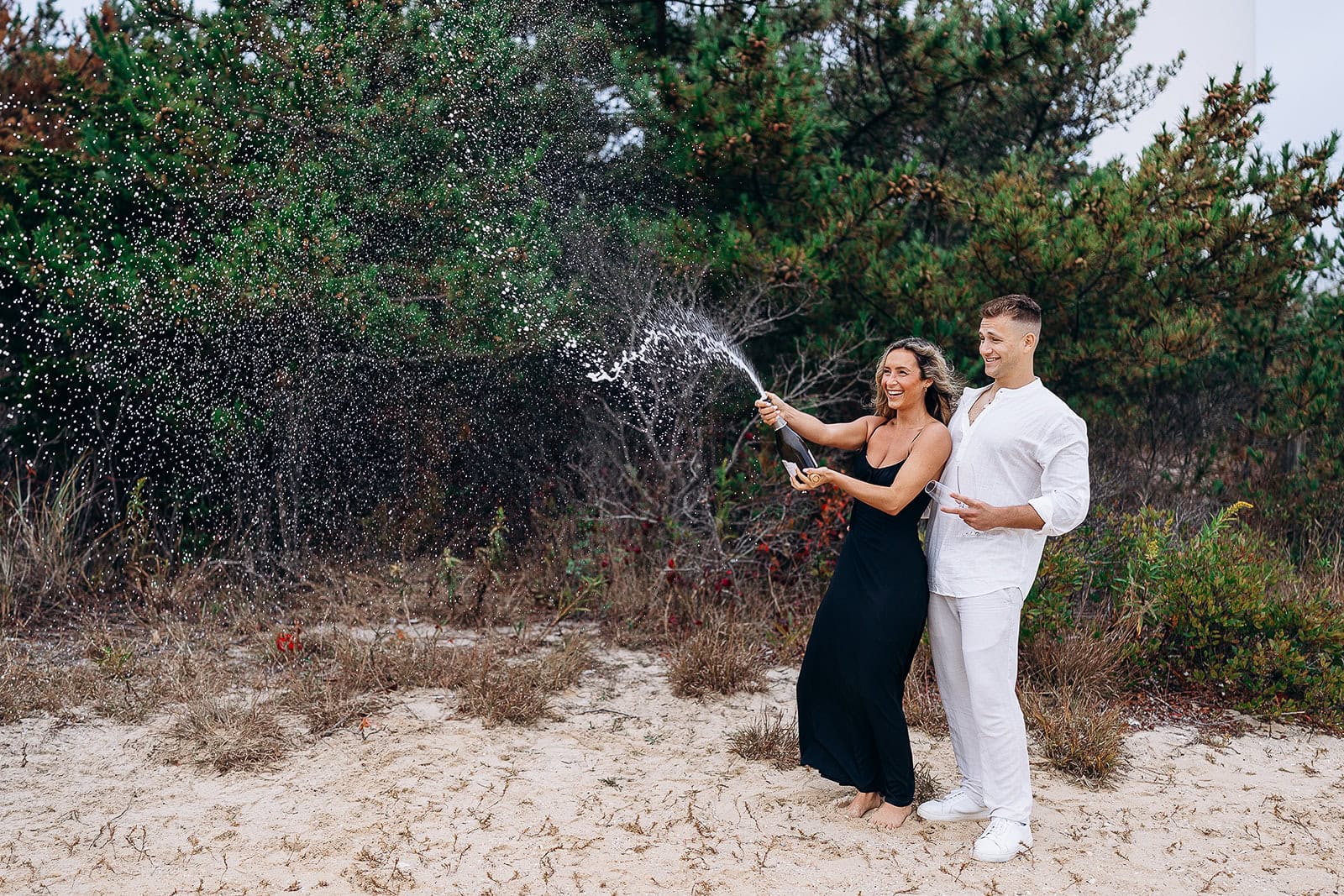 Couple on sandy beach with dense green foliage behind, woman in black dress spraying champagne, man in white outfit holding glass