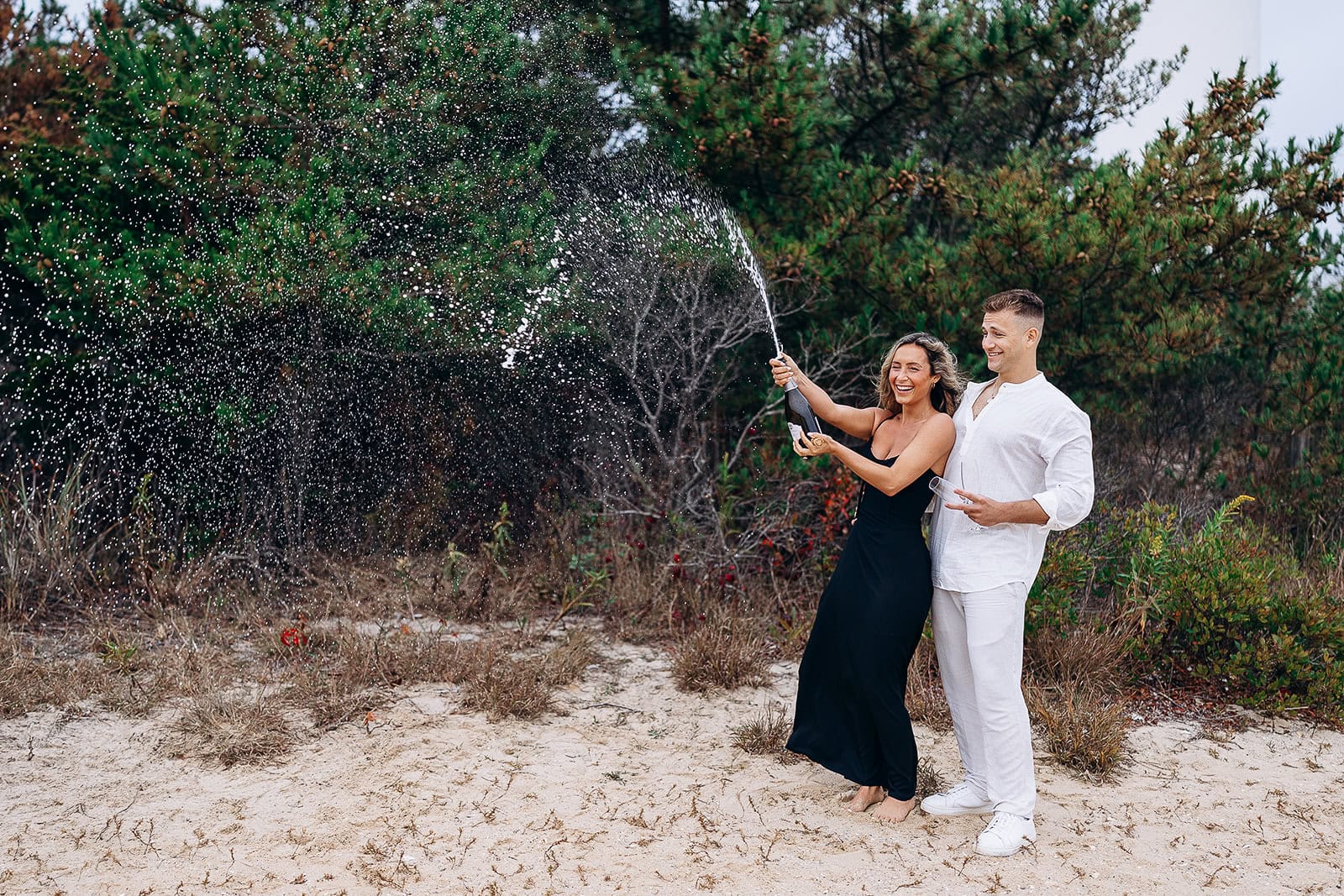 Couple standing barefoot on sandy ground near green pine trees, woman in black dress popping champagne bottle, man in white shirt holding glass, Barnegat Light, NJ.
