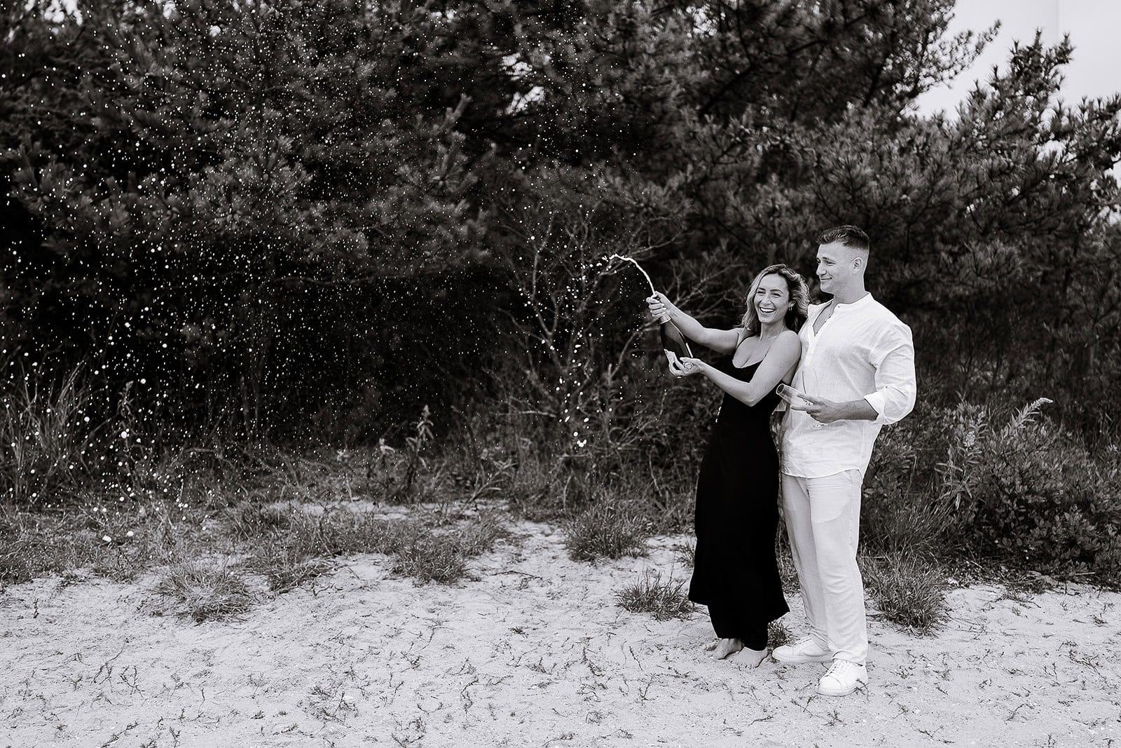 Couple standing on sandy ground near dense trees, woman in black dress popping champagne, man in white shirt holding a glass, champagne spray in air