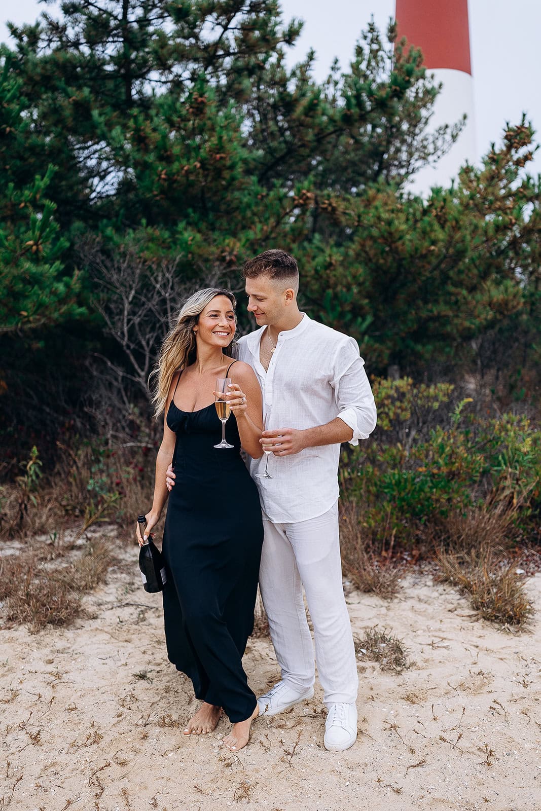 Couple standing barefoot on sandy ground near Barnegat Light lighthouse, holding champagne glasses and a bottle, surrounded by green foliage.