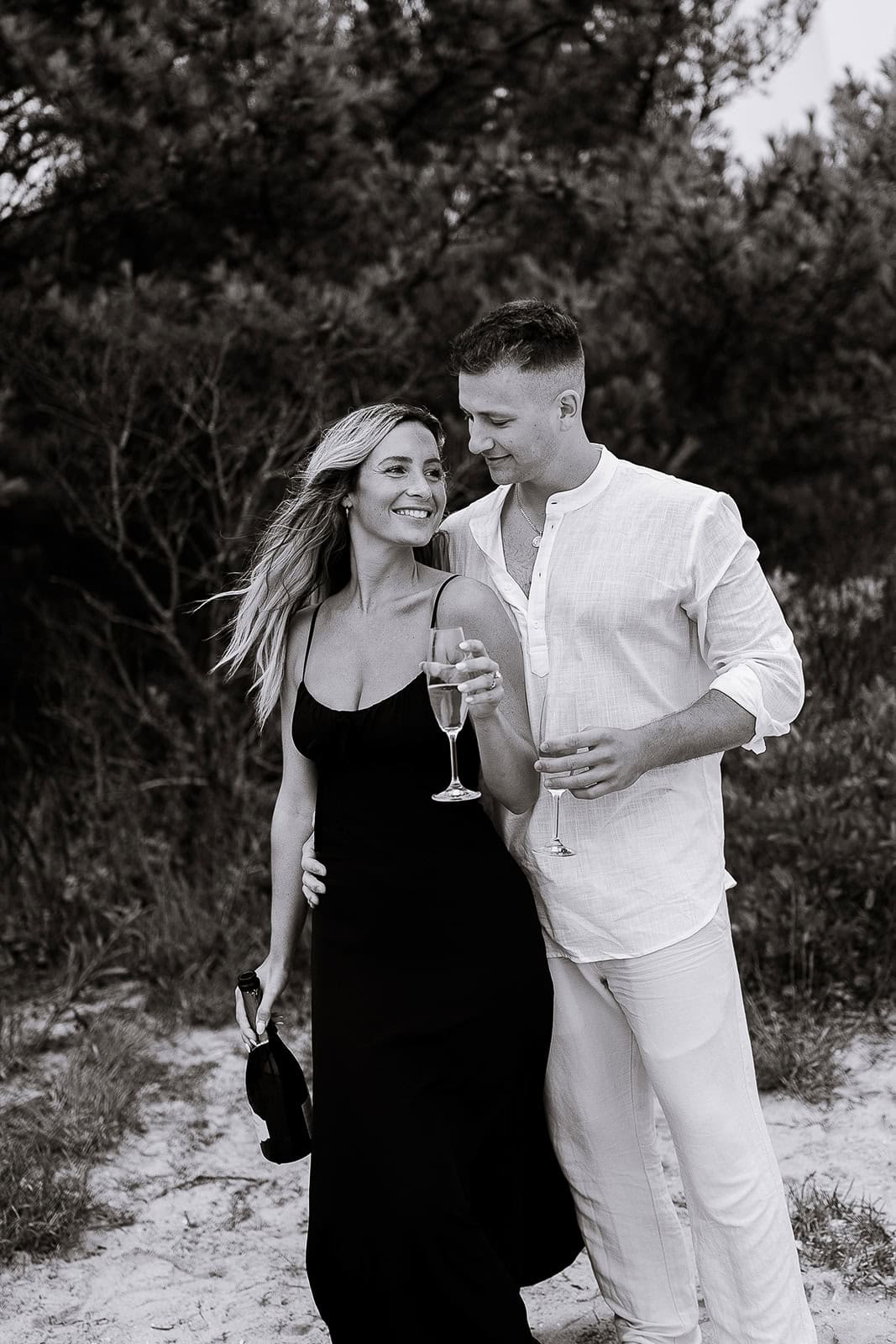 Black and white image of a couple standing closely outdoors, woman in a black dress holding a champagne bottle and glass, man in white shirt and pants holding a glass, surrounded by trees.