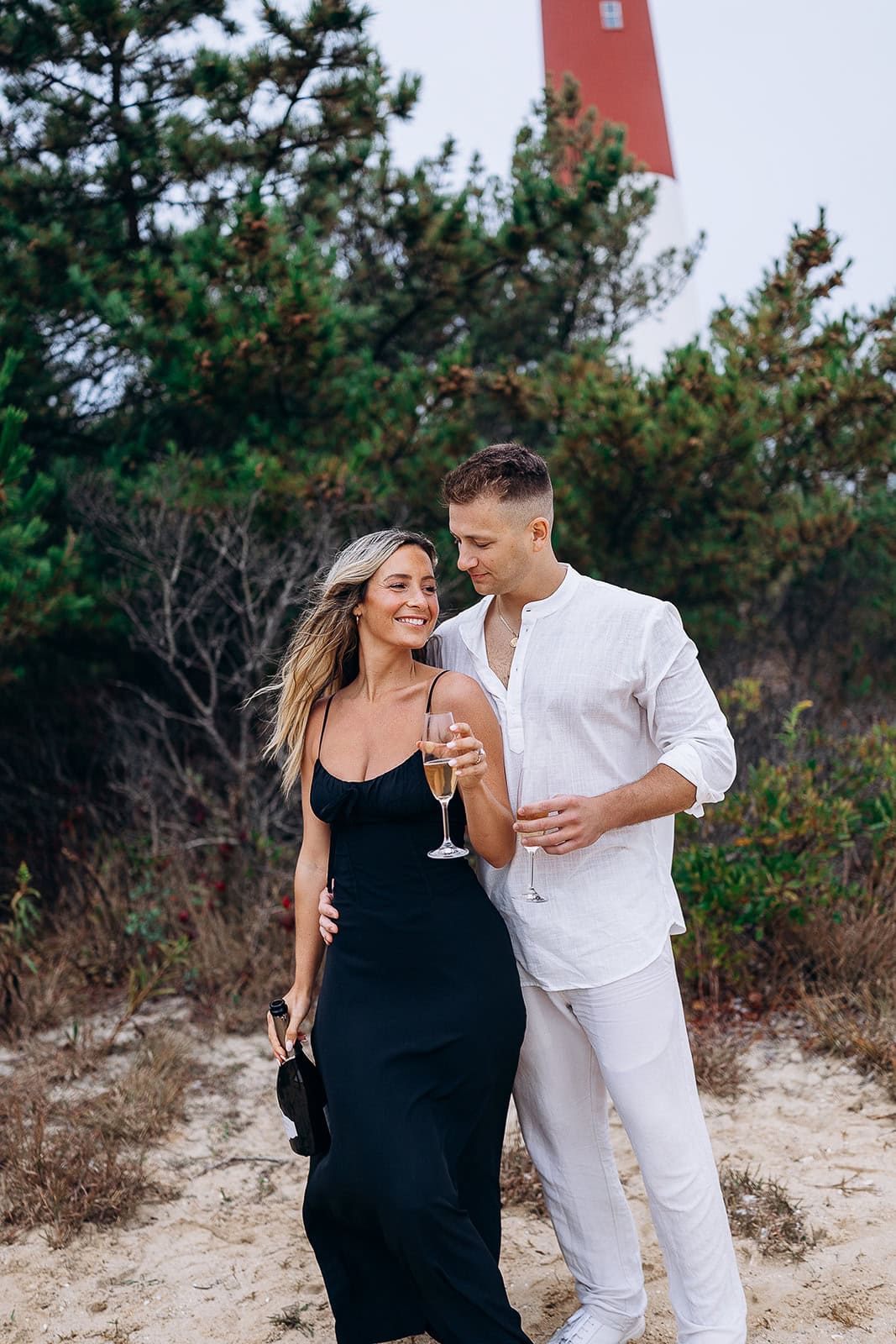 Couple standing on sandy ground near green shrubs with Barnegat Light lighthouse in background, holding champagne glasses and bottle.