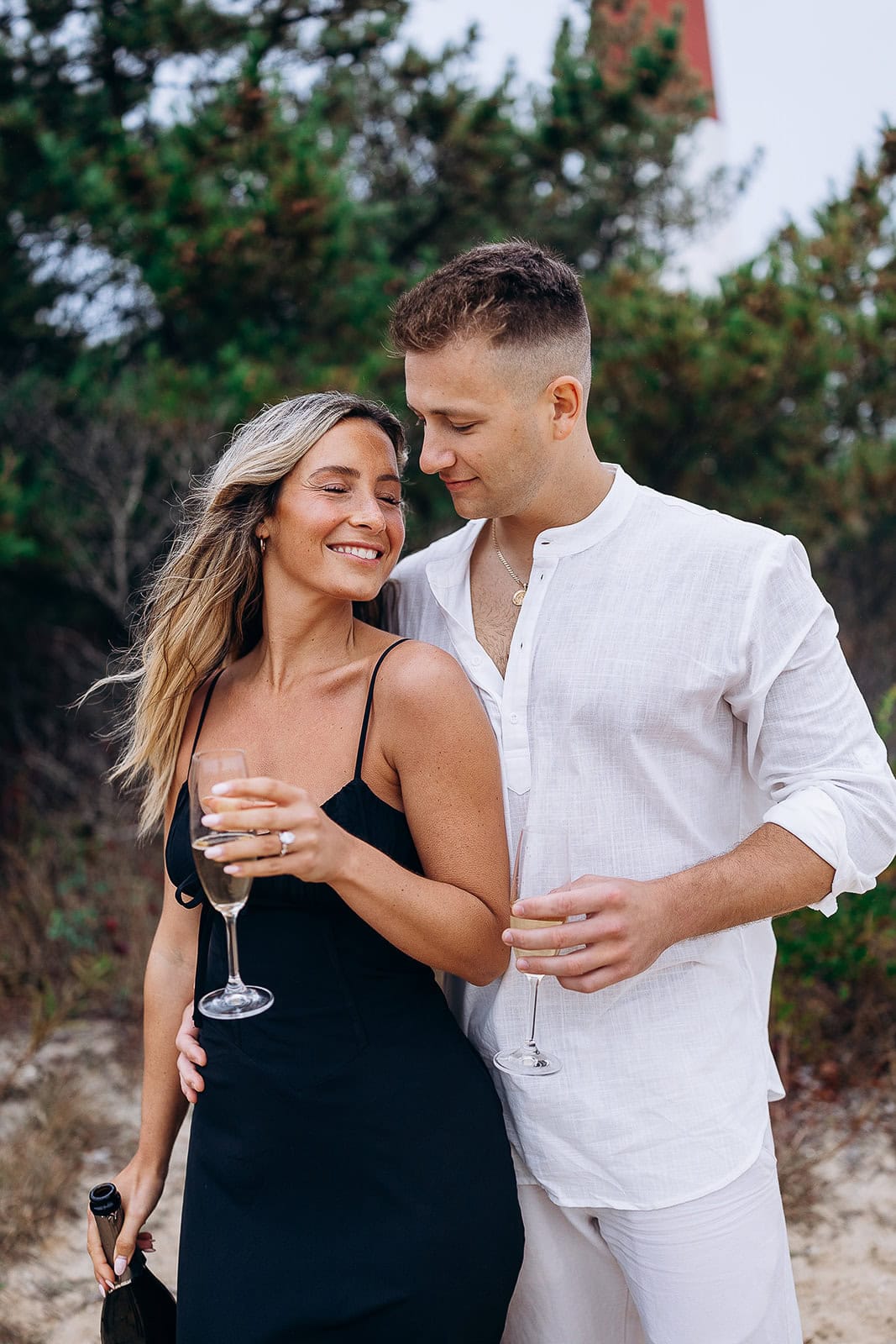 Couple in sophisticated attire holding champagne glasses outdoors near greenery, sharing an intimate moment.