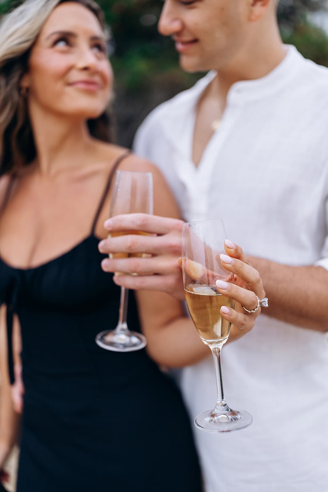 Close-up of a couple holding champagne flutes, the woman in a black dress and the man in a white shirt, softly smiling at each other.