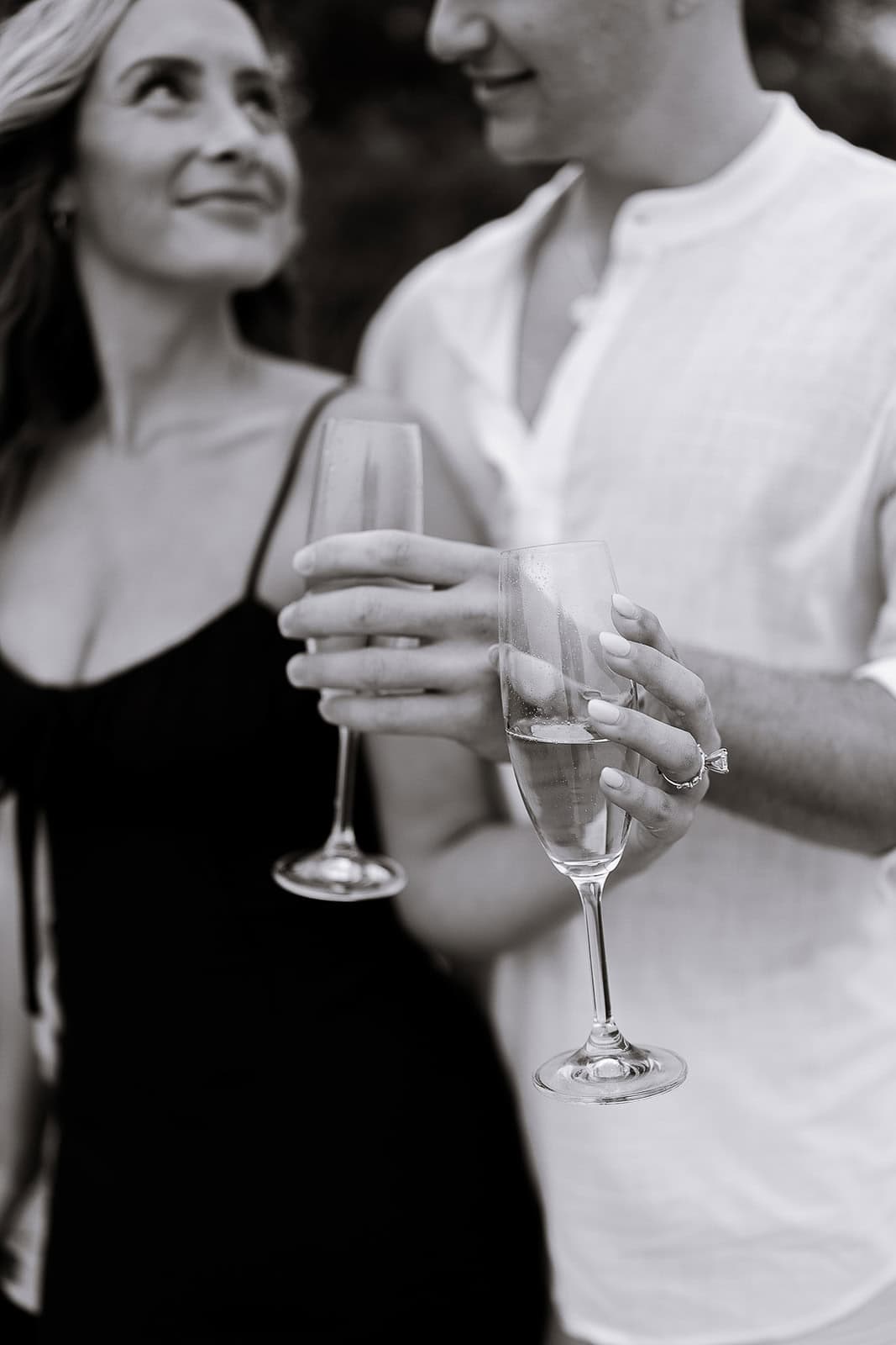 Close-up of a couple holding champagne flutes, woman in black dress with engagement ring, man in white shirt, soft focus background.