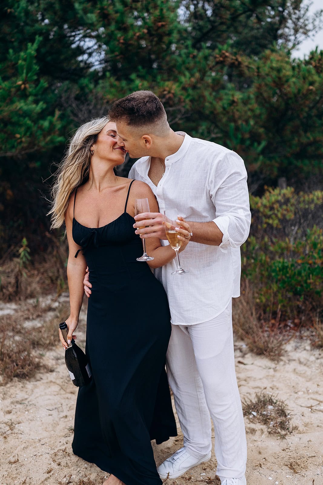 Couple sharing a tender kiss outdoors surrounded by greenery, woman in black dress holding a champagne bottle, man in white shirt holding two champagne glasses.
