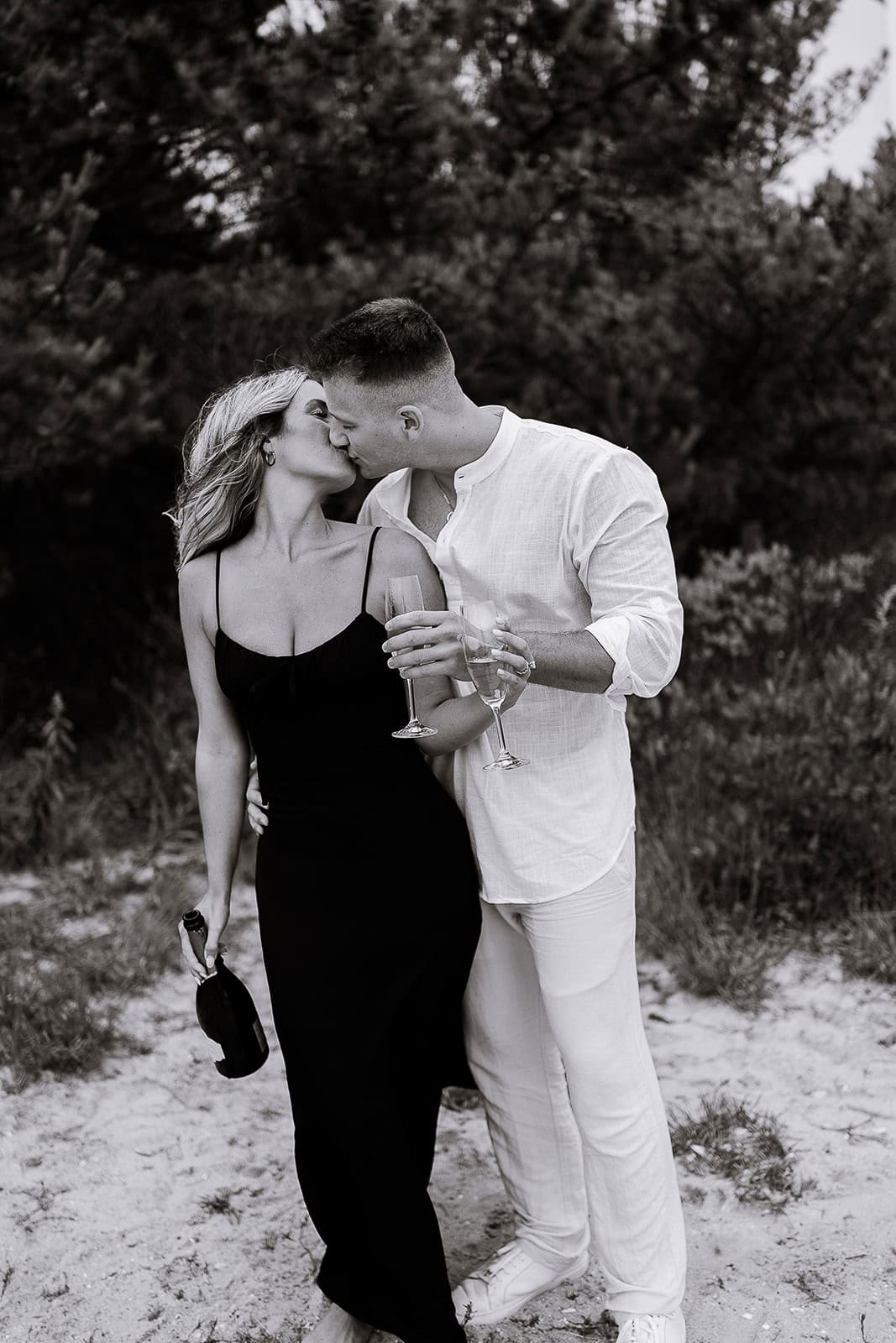 Couple kissing outdoors on sandy ground near trees, holding champagne glasses and bottle, in Barnegat Light.