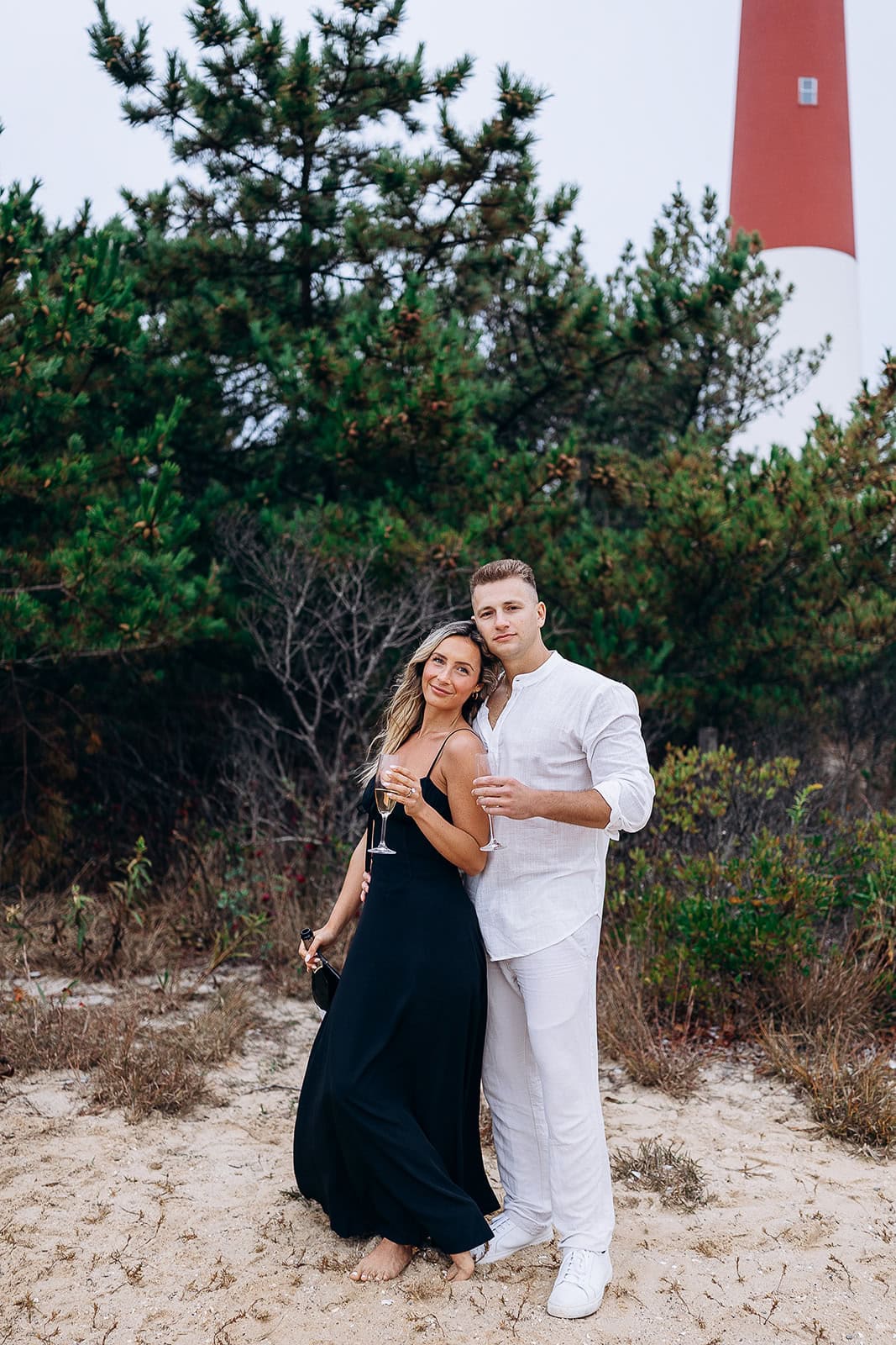 Couple standing barefoot on sandy ground near pine trees, holding champagne glasses, with Barnegat Light lighthouse in the background.