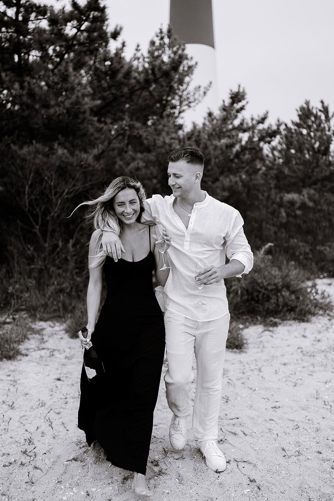Couple walking barefoot on sandy beach at Barnegat Light, NJ, holding champagne glasses and bottle, with lighthouse and trees in background.