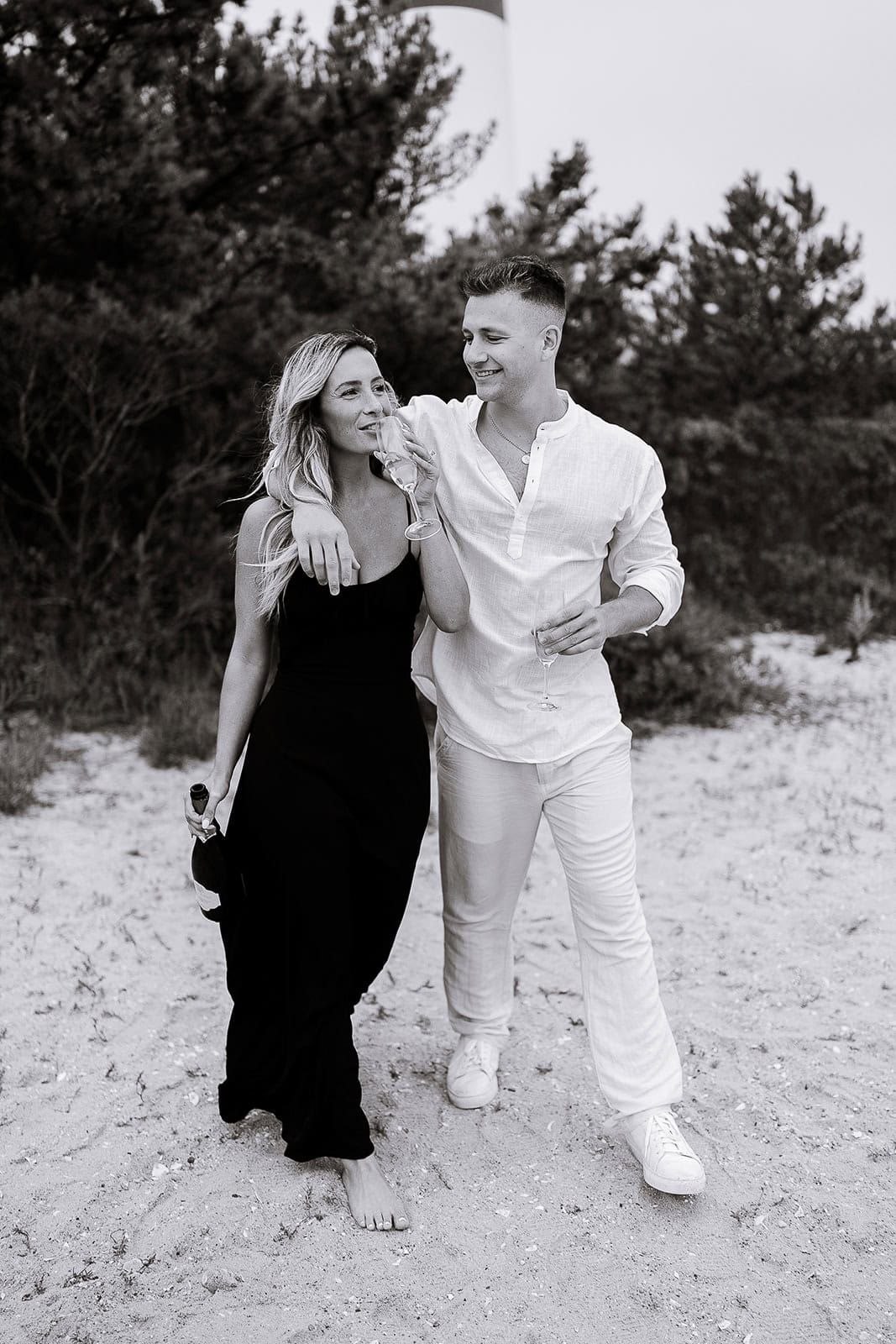Black and white image of a couple walking barefoot on sandy ground with trees in the background; the woman carries a bottle and drinks from a glass while the man has his arm around her shoulder holding a glass.