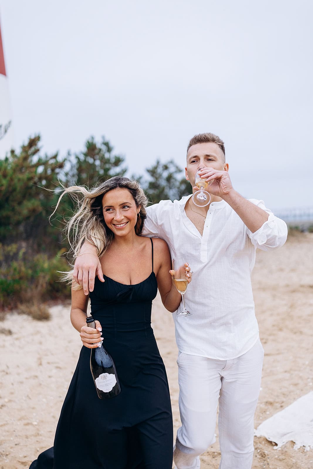 Couple walking on a sandy path in Barnegat Light, NJ; woman in black dress holding a champagne bottle, man in white shirt and pants drinking from a glass.