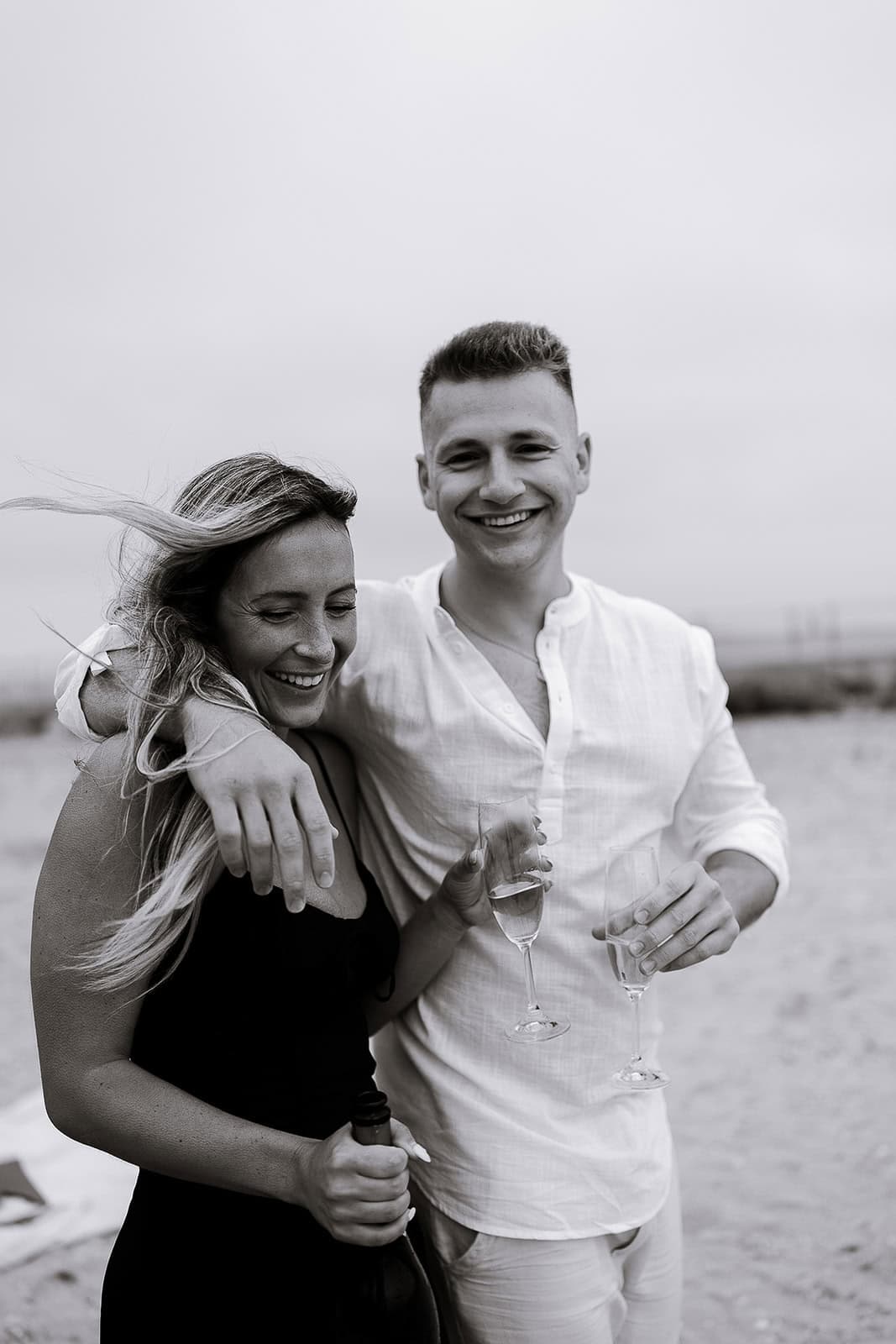 Black and white photo of a smiling couple on a beach, the man in a white shirt with his arm around the woman in a black dress holding a bottle and champagne glasses.