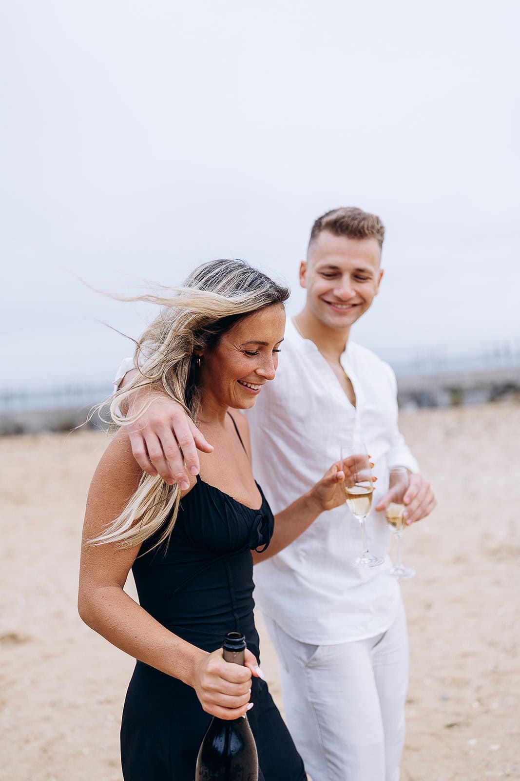 A man in white linen and a woman in a black dress walk arm in arm on a sandy beach, holding champagne glasses and a bottle in a breezy setting.