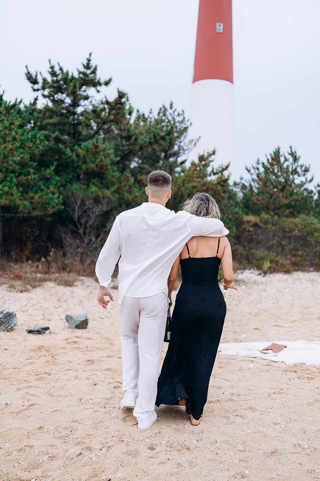 Couple walking arm in arm on sandy beach with Barnegat Light lighthouse and pine trees in the background.