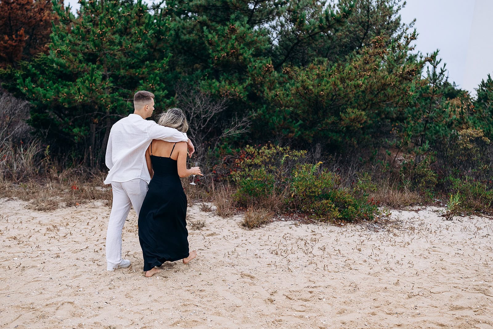 Couple walking barefoot on sandy beach near dense green foliage in Barnegat Light, NJ, the man in white and the woman in a black dress holding a glass.