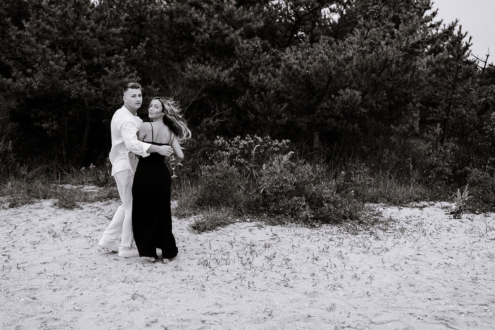 Black and white image of a man in white and a woman in a black dress embracing on a sandy beach with dense trees in the background.