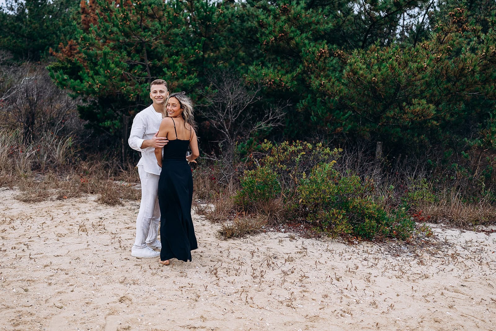 Couple standing on sandy ground near dense green shrubs and trees, the man in white attire gently holding the woman in a black dress, outdoors in a natural coastal setting.