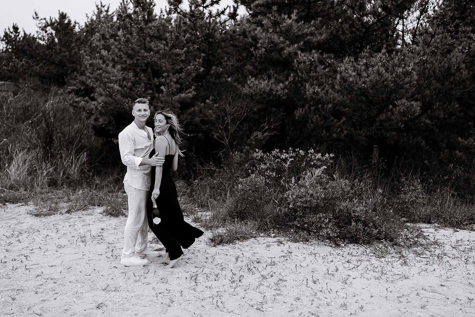 Black and white image of a couple standing barefoot on a sandy beach with dense pine trees in the background, the man in light clothing and the woman in a flowing dark dress.