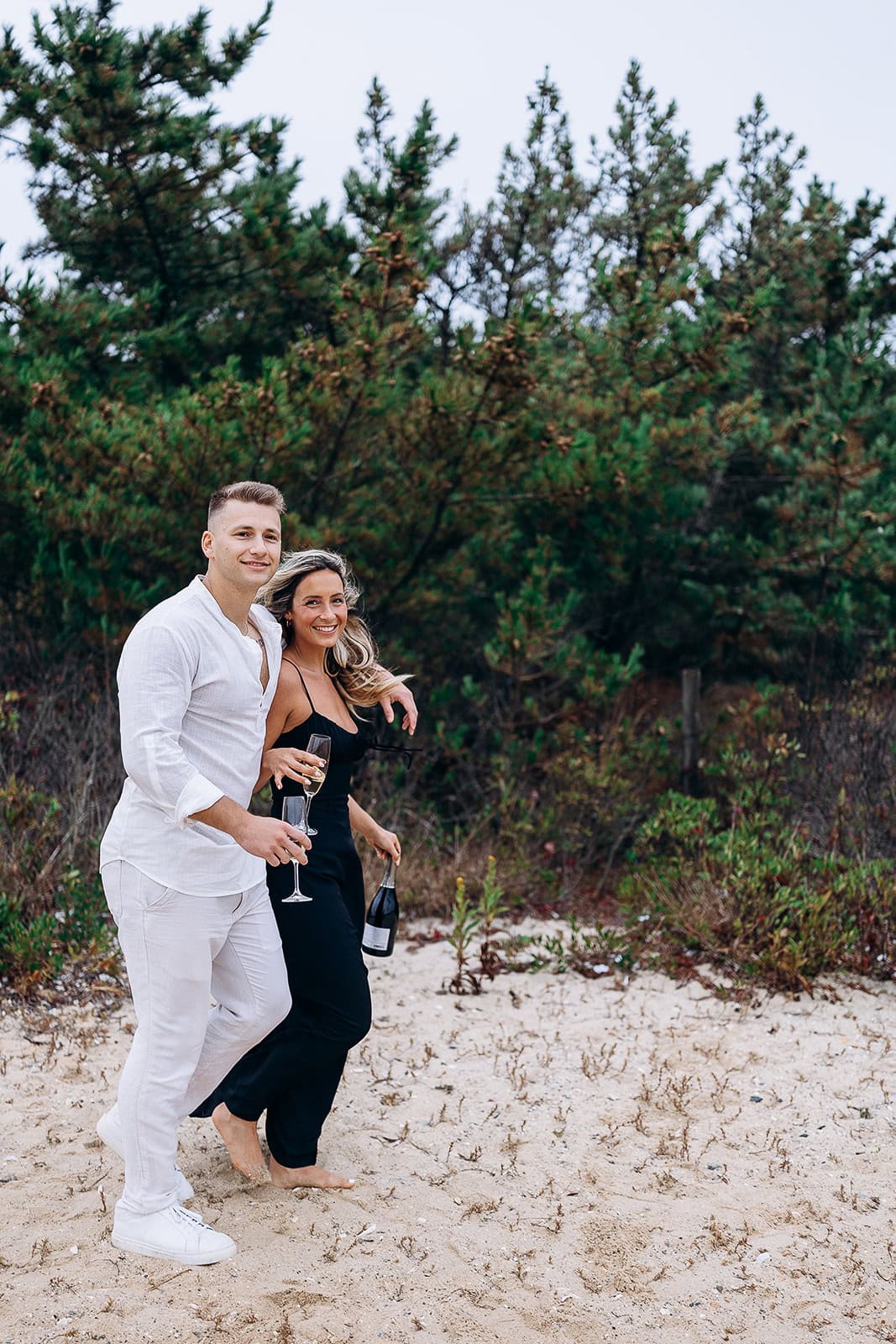 Man in white linen shirt and pants walking barefoot with woman in black jumpsuit holding champagne bottle and glasses on sandy path near green pine trees in Barnegat Light
