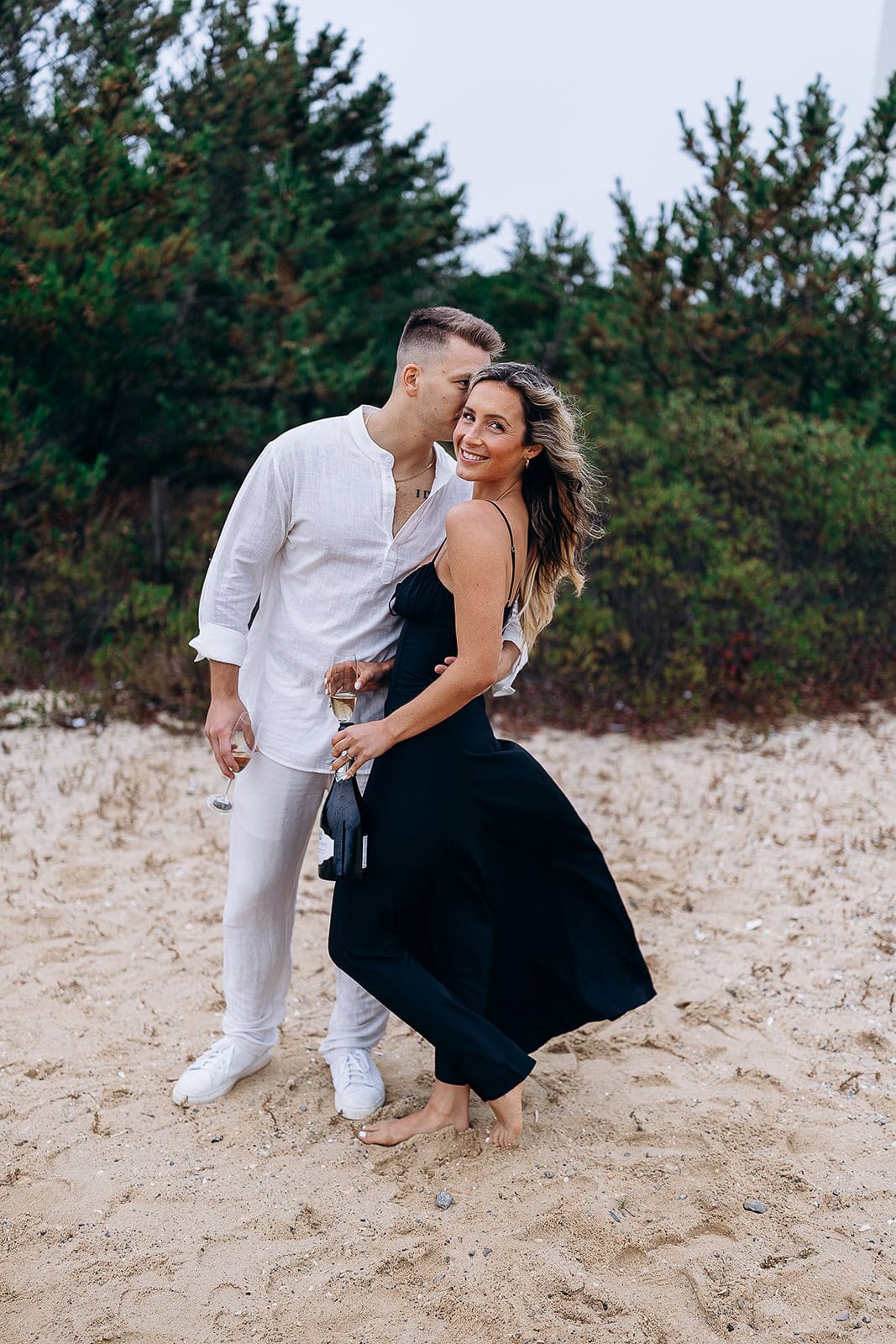 Couple on sandy beach with pine trees in background; man in white linen outfit kisses woman in black dress holding champagne bottle and glass.