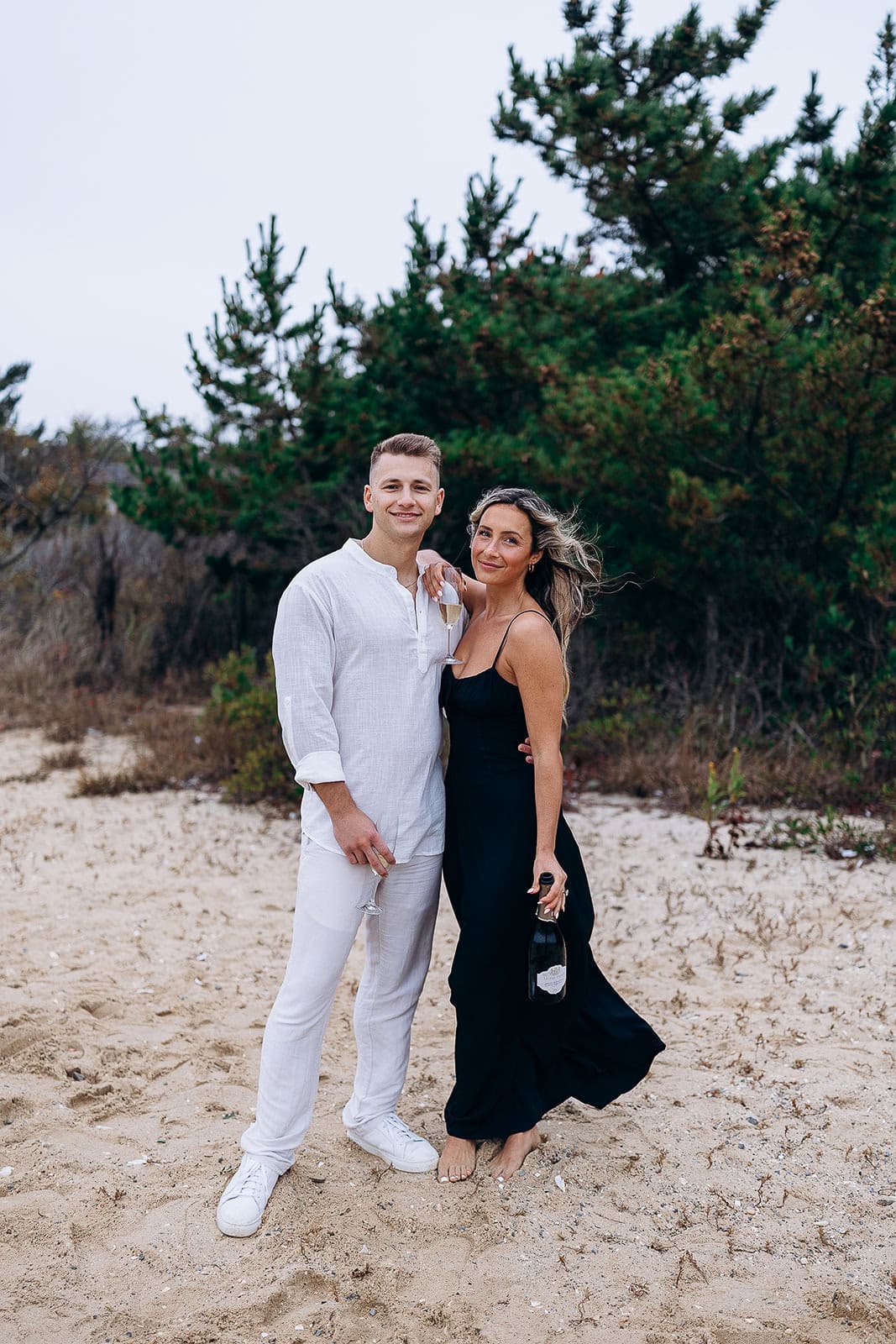 Couple standing on sandy beach with pine trees behind; woman in black dress holding champagne bottle, man in white outfit holding glass
