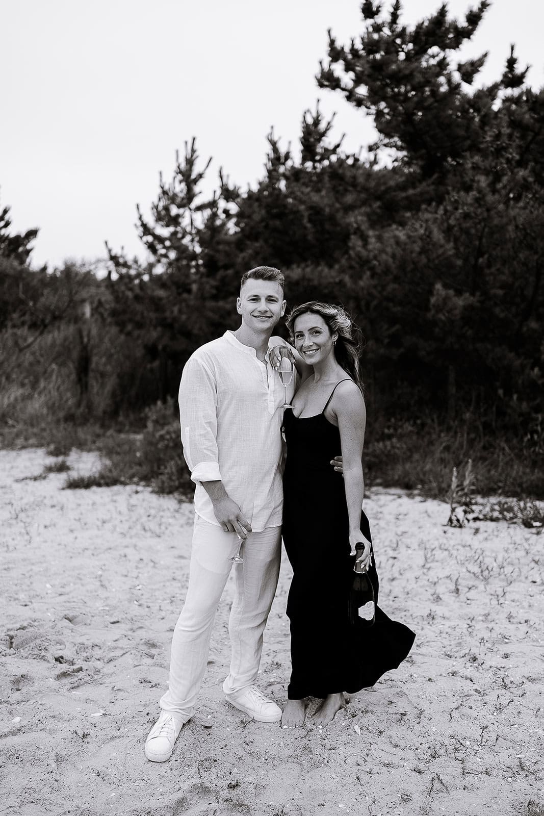 A couple stands barefoot on sandy ground with trees behind them; the man wears white linen and sneakers, the woman a black dress holding a champagne bottle and glass.
