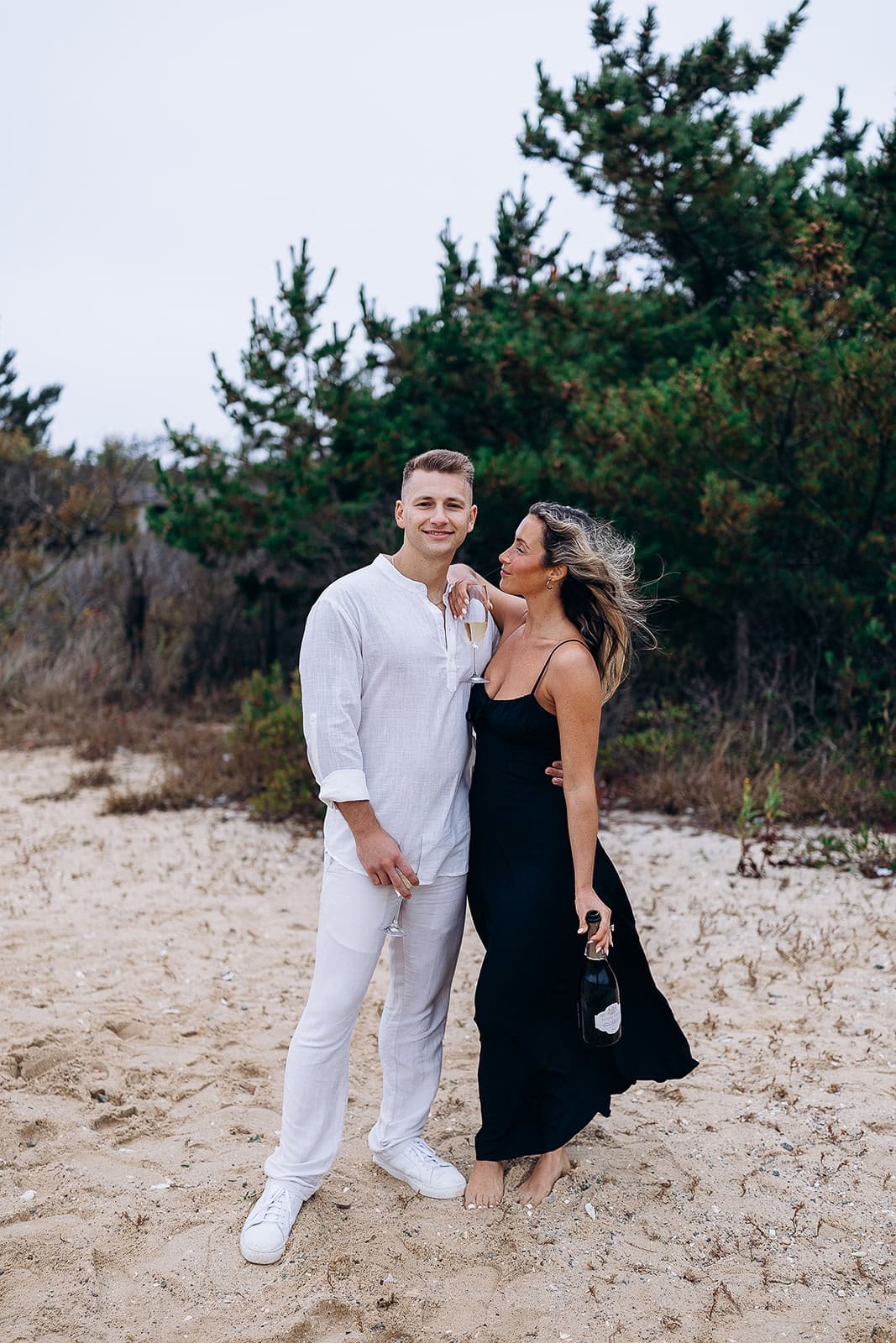 Couple on sandy beach with greenery background; woman in black dress holding champagne bottle, man in white outfit holding champagne flute.