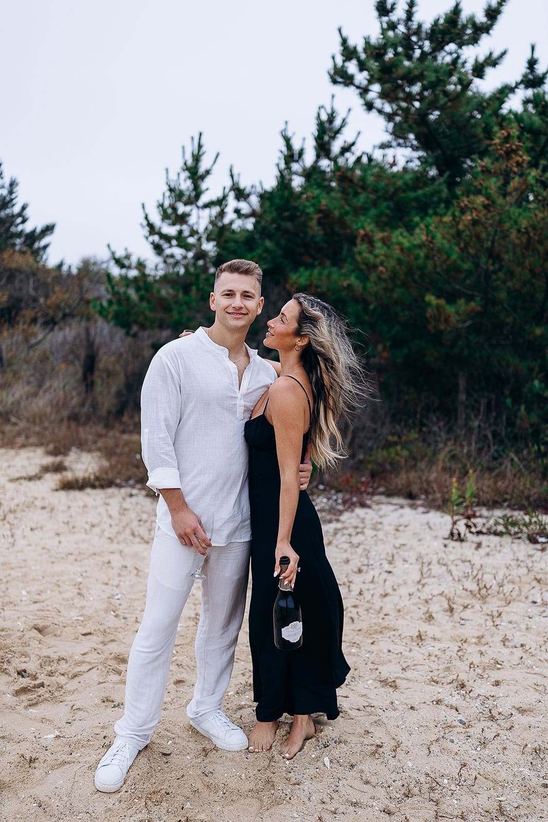 Couple standing barefoot on sandy beach near pine trees, man in white linen outfit holding a glass, woman in black dress holding a bottle.