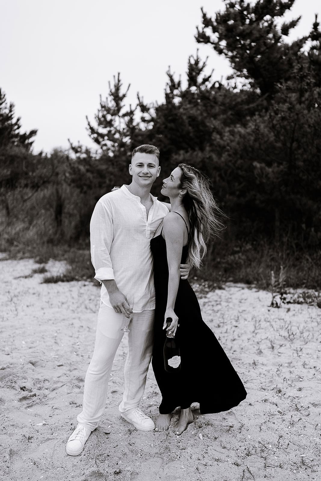 Black and white photo of a couple standing barefoot on a sandy beach with trees in the background. The man wears white linen clothing and holds a wine glass, while the woman in a flowing black dress holds a bottle of champagne.