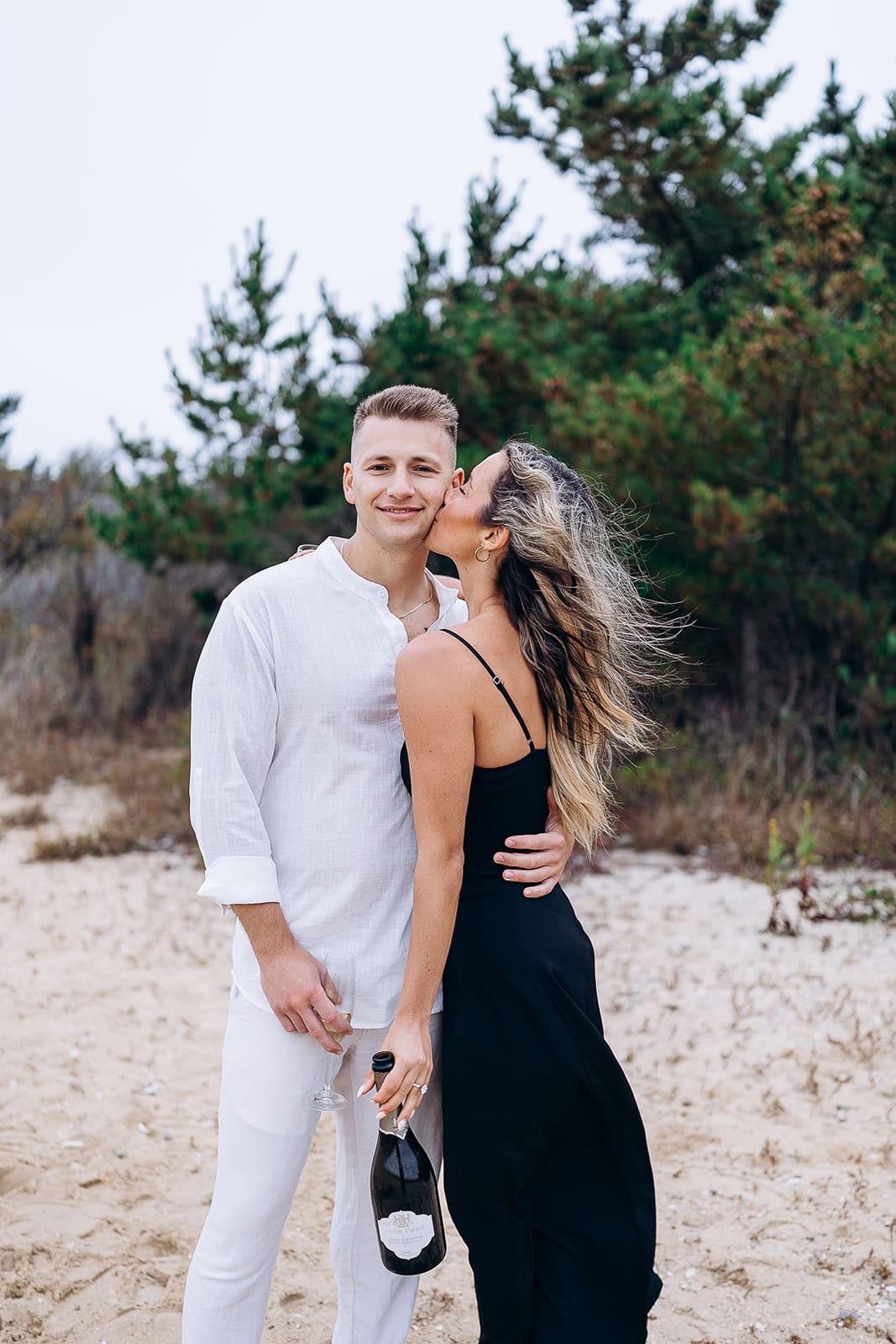 Couple on sandy beach with pine trees in background; woman in black dress kisses man in white shirt holding champagne bottle and glass.