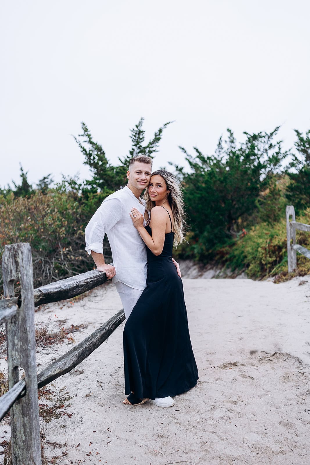 Couple standing close on sandy path at Barnegat Light, NJ, with rustic wooden fence and green foliage in background.