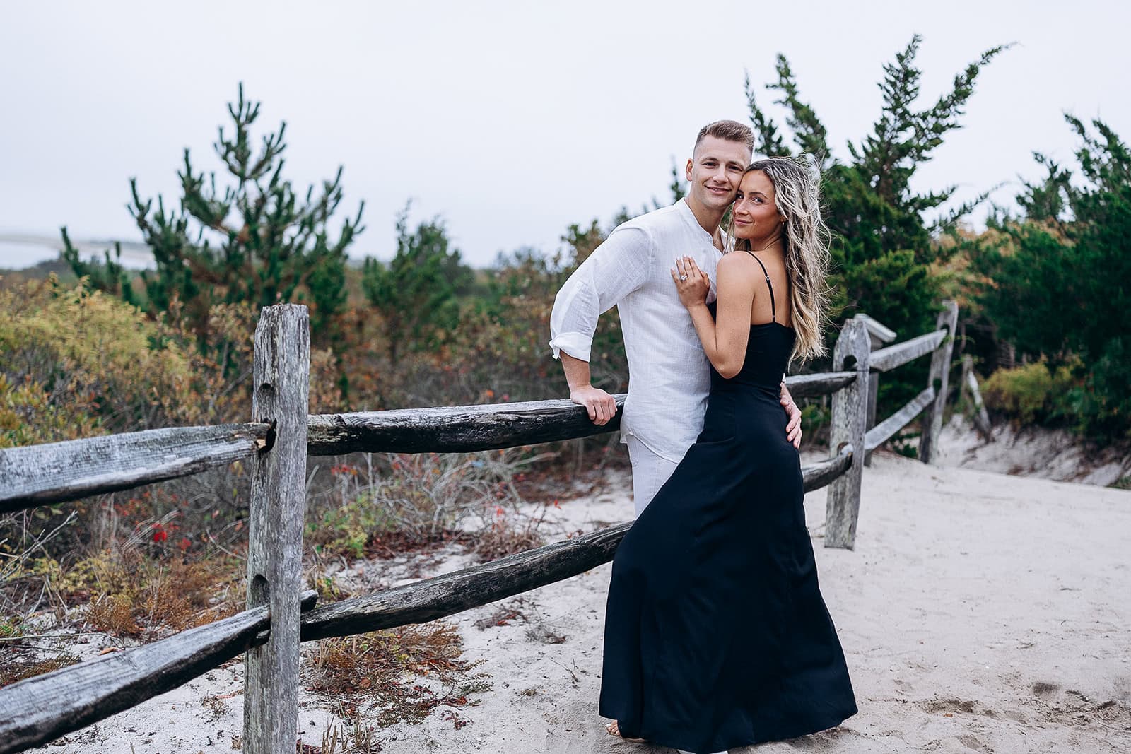 Couple embracing near rustic wooden fence on sandy path with coastal greenery at Barnegat Light, New Jersey.