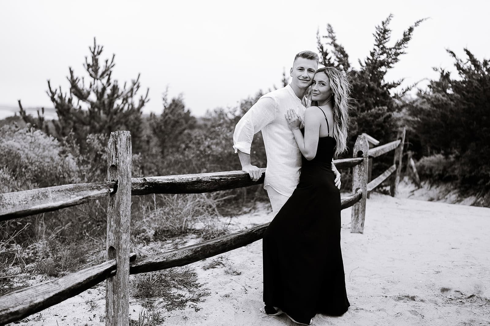 Black and white photo of a couple standing by a weathered wooden fence on a sandy path, surrounded by pine trees and shrubbery.