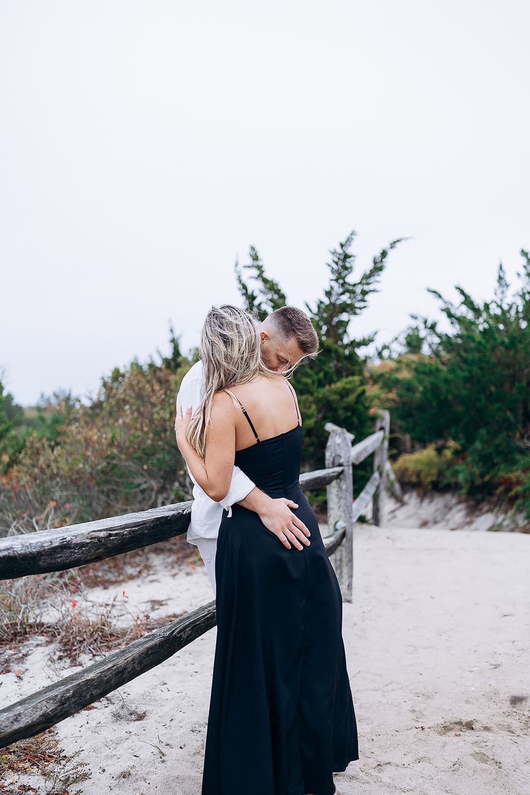 Couple embracing near rustic wooden fence on sandy path with greenery in Barnegat Light, NJ.