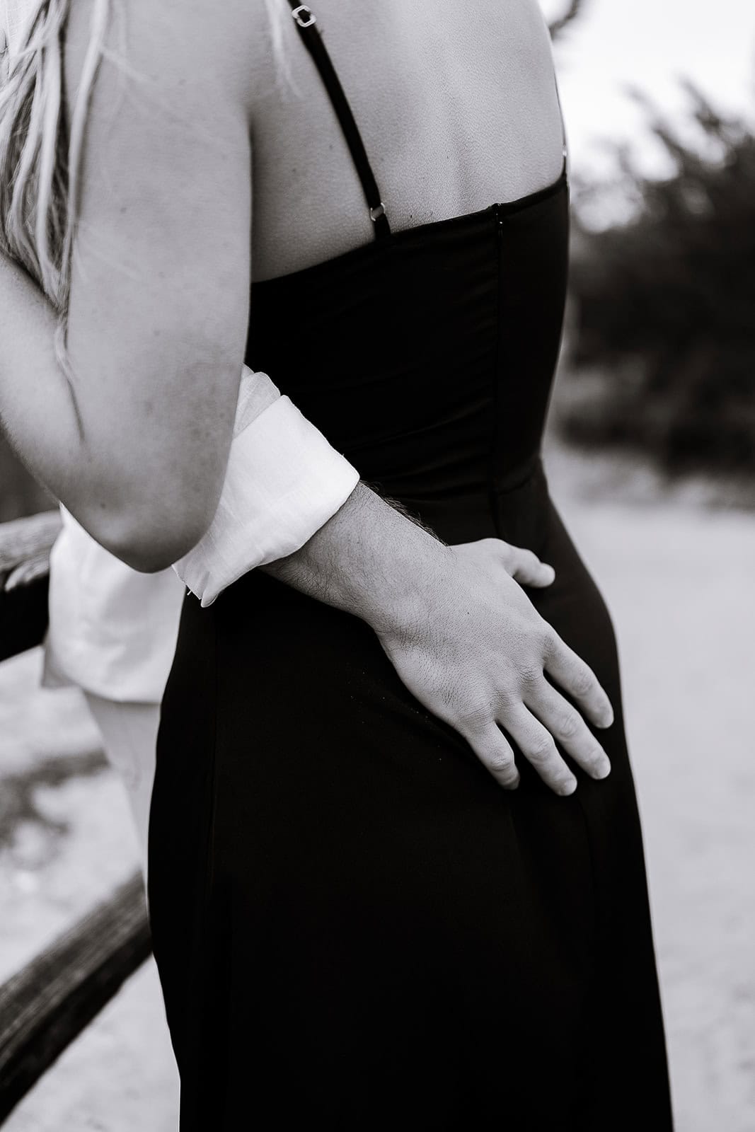 Close-up black and white photo of a man’s hand resting gently on a woman’s waist, the woman wearing a black dress with thin straps, set against a blurred outdoor background.