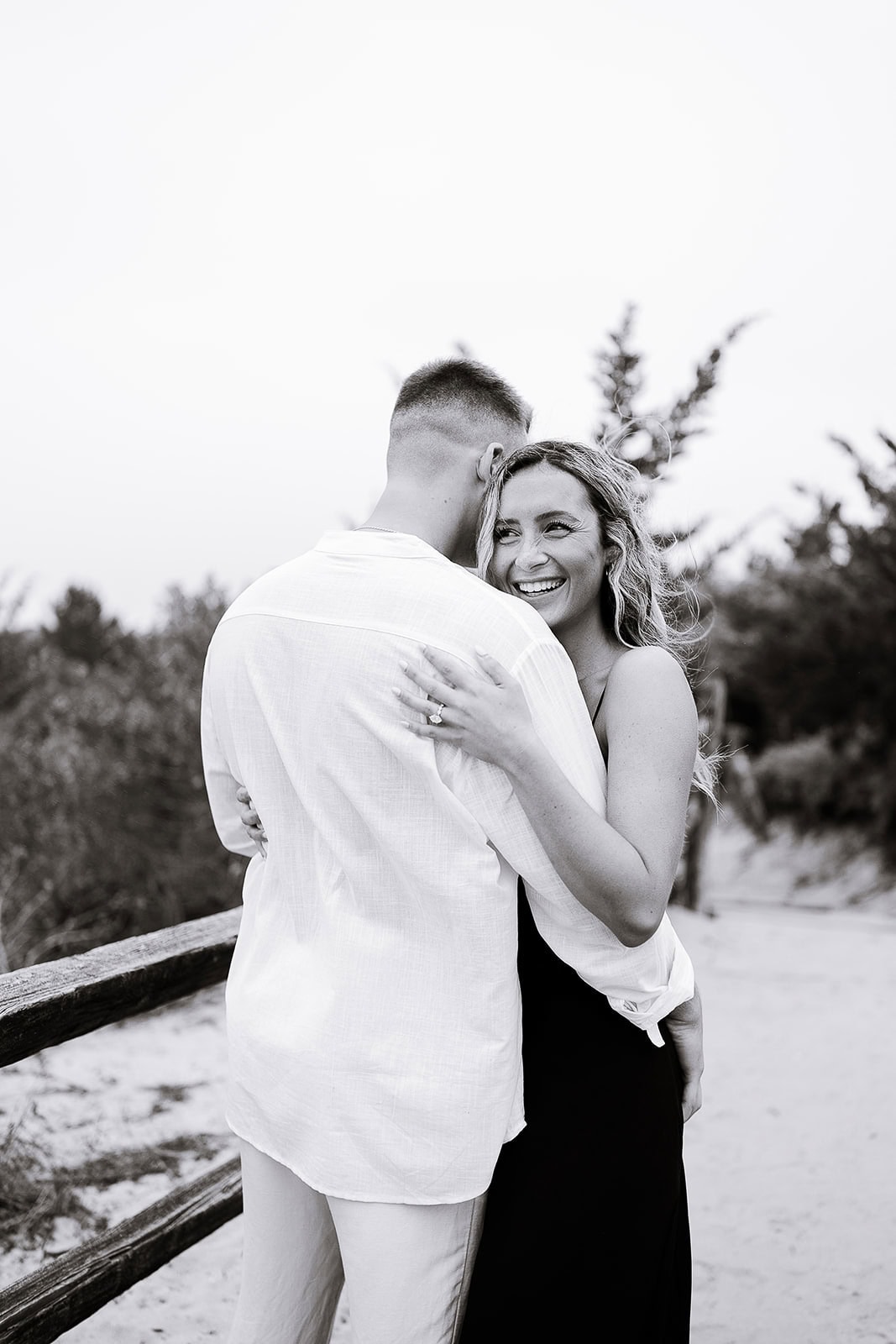 Black and white image of a couple embracing outdoors by a wooden fence, the woman smiling warmly while holding the man, surrounded by trees and sand.