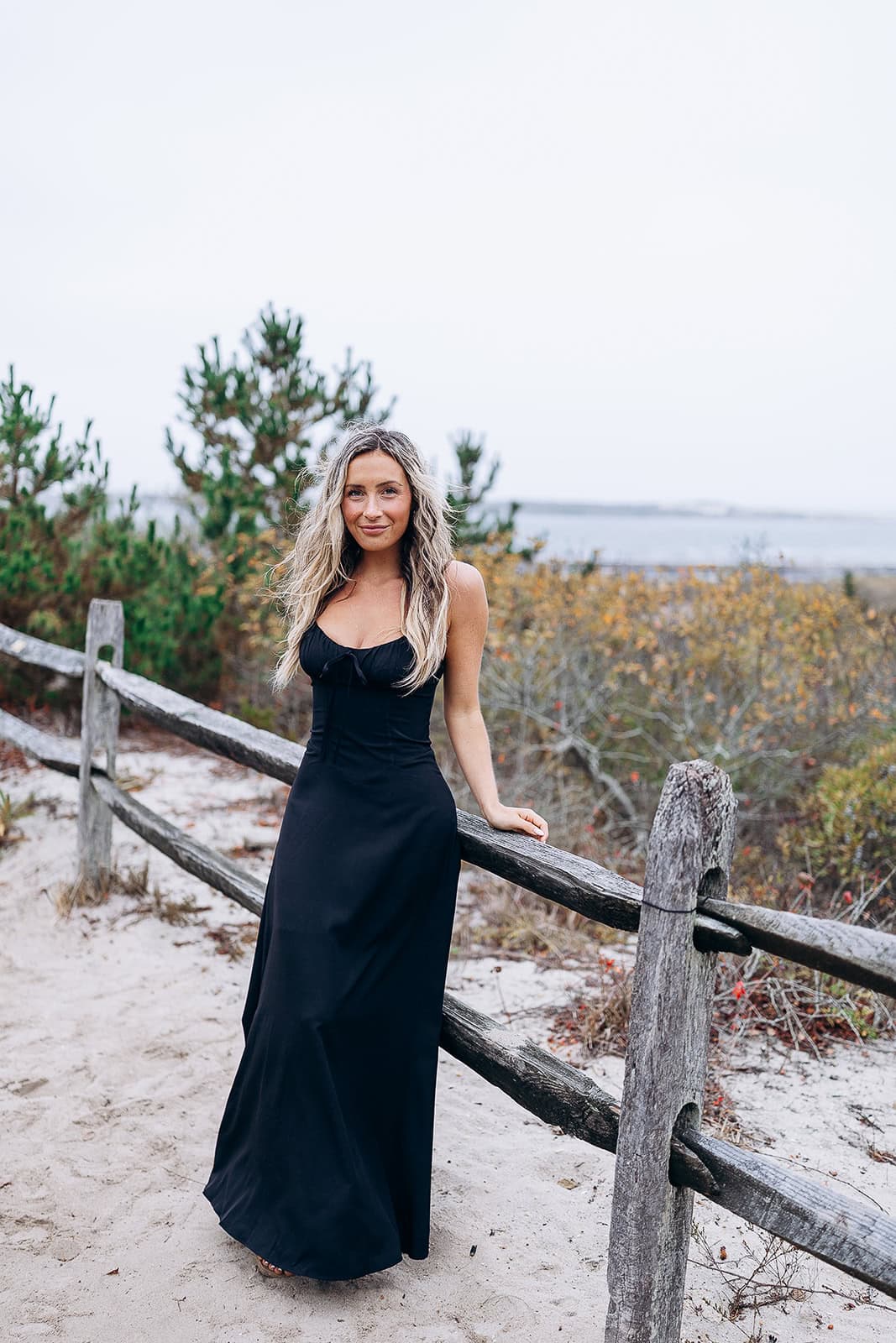 Woman in a long black dress standing by a rustic wooden fence on sandy ground with coastal shrubs and water in the background at Barnegat Light.