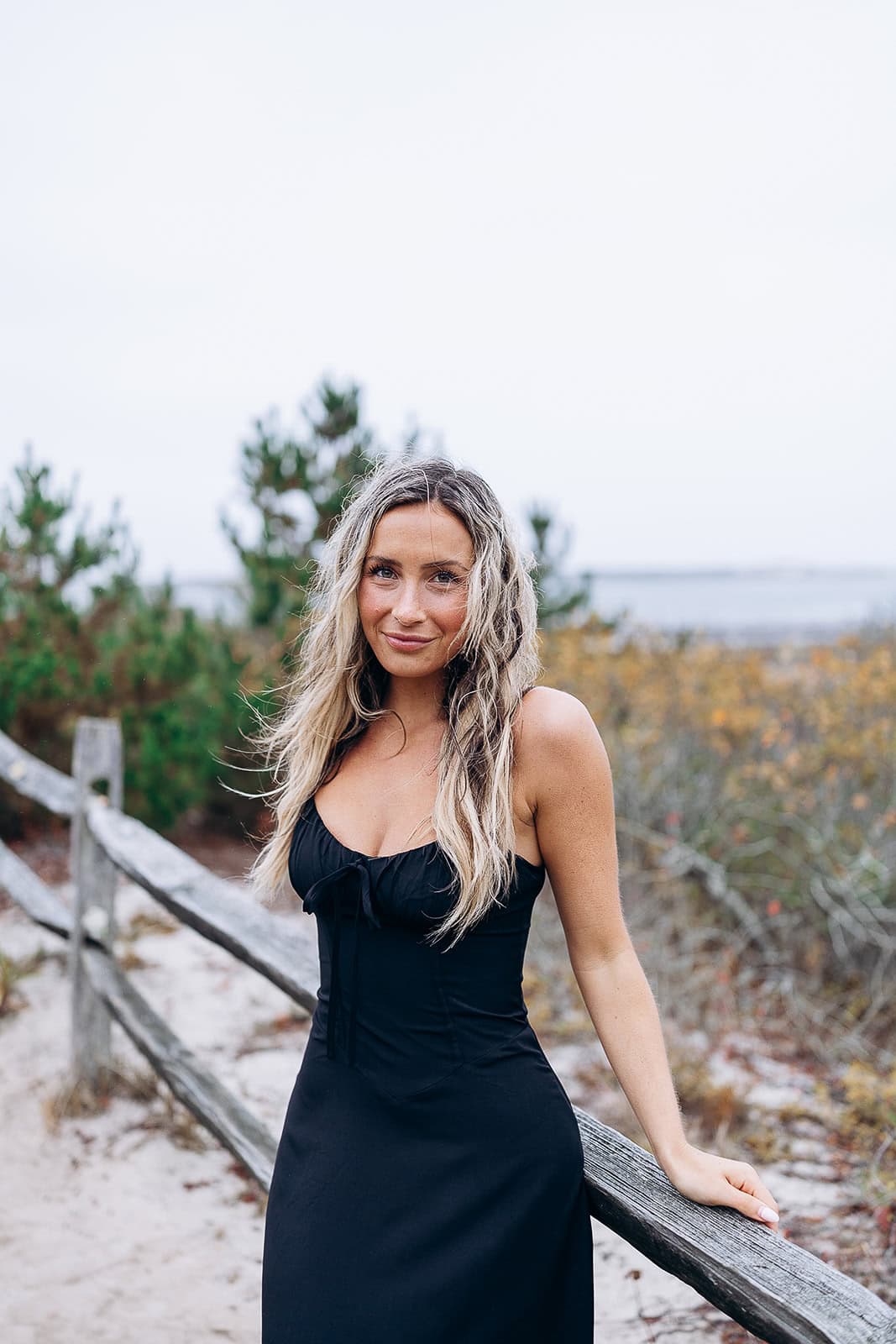 Woman with long wavy blonde hair wearing a black dress leaning on a rustic wooden fence with dunes and autumn foliage in Barnegat Light.