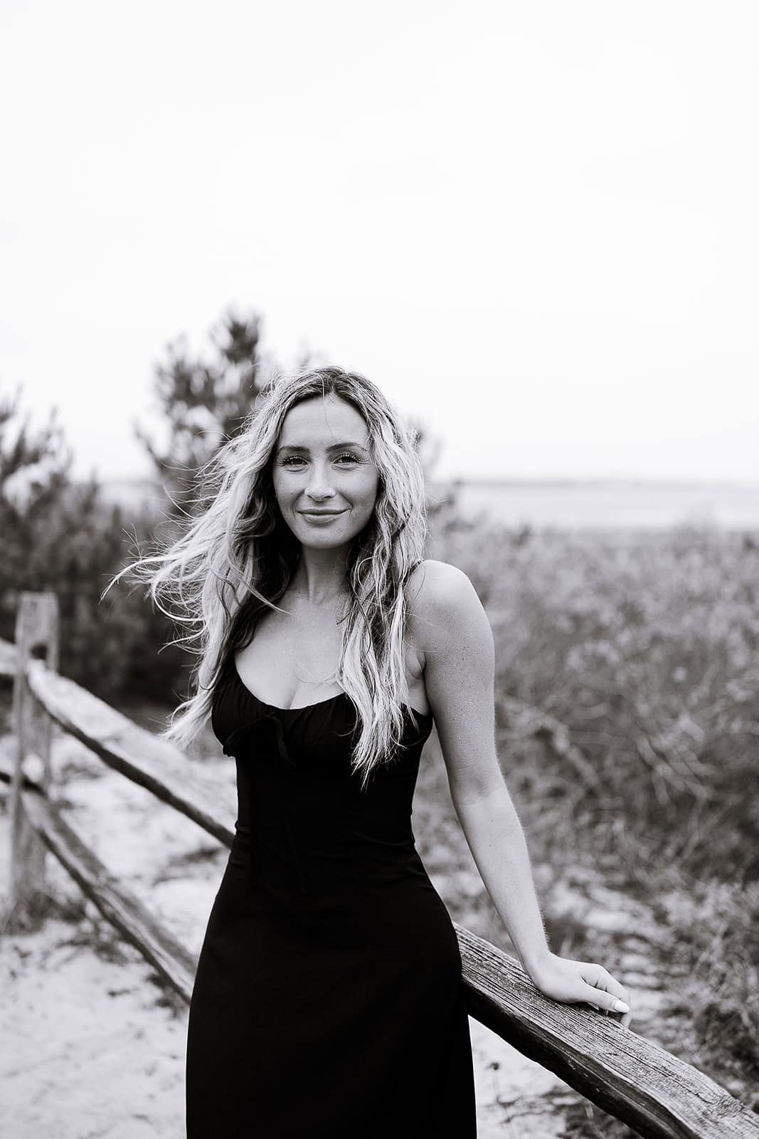 Black-and-white portrait of a woman in a black dress leaning on a wooden fence with sand dunes and sparse trees in the background at Barnegat Light, NJ.
