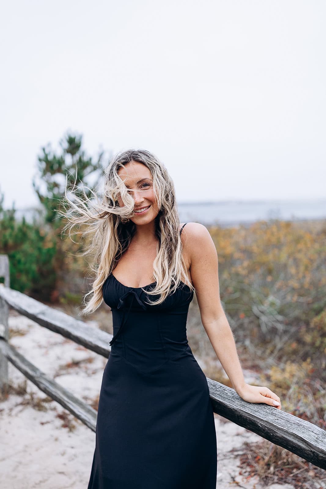 Woman in a black dress leaning on a weathered wooden fence with windswept hair, coastal dunes and sparse vegetation in Barnegat Light.