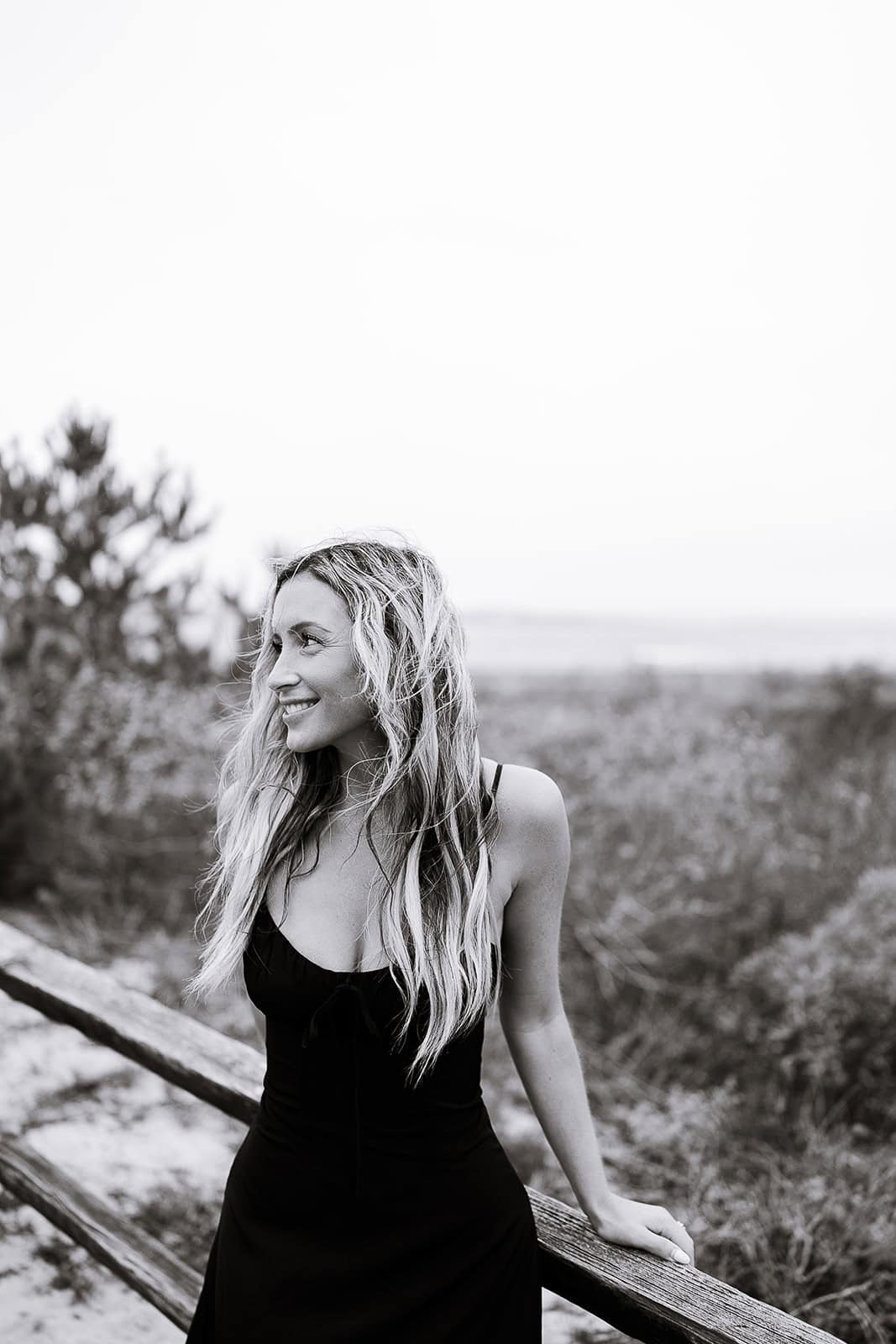 Black and white image of a woman with long wavy hair in a black dress leaning on a wooden railing, looking to the left with a blurred natural landscape behind, Barnegat Light, NJ.