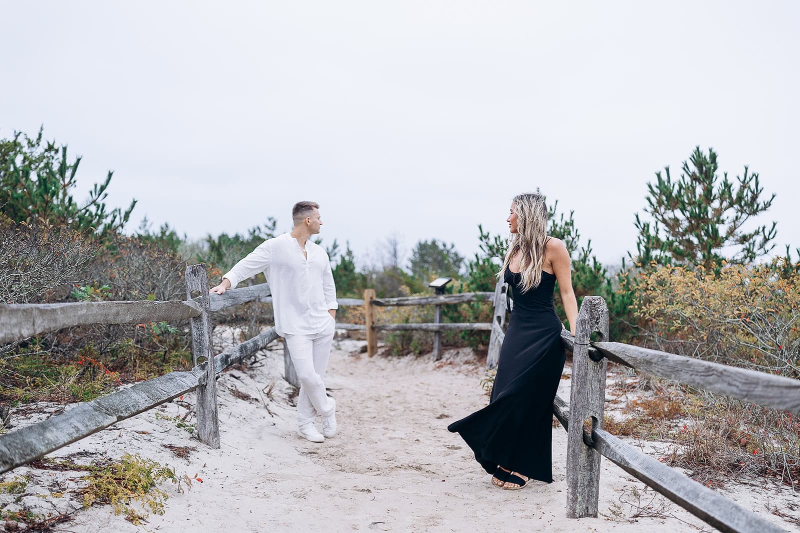 Couple standing apart on sandy path with weathered wooden fence and coastal shrubs in Barnegat Light, NJ.