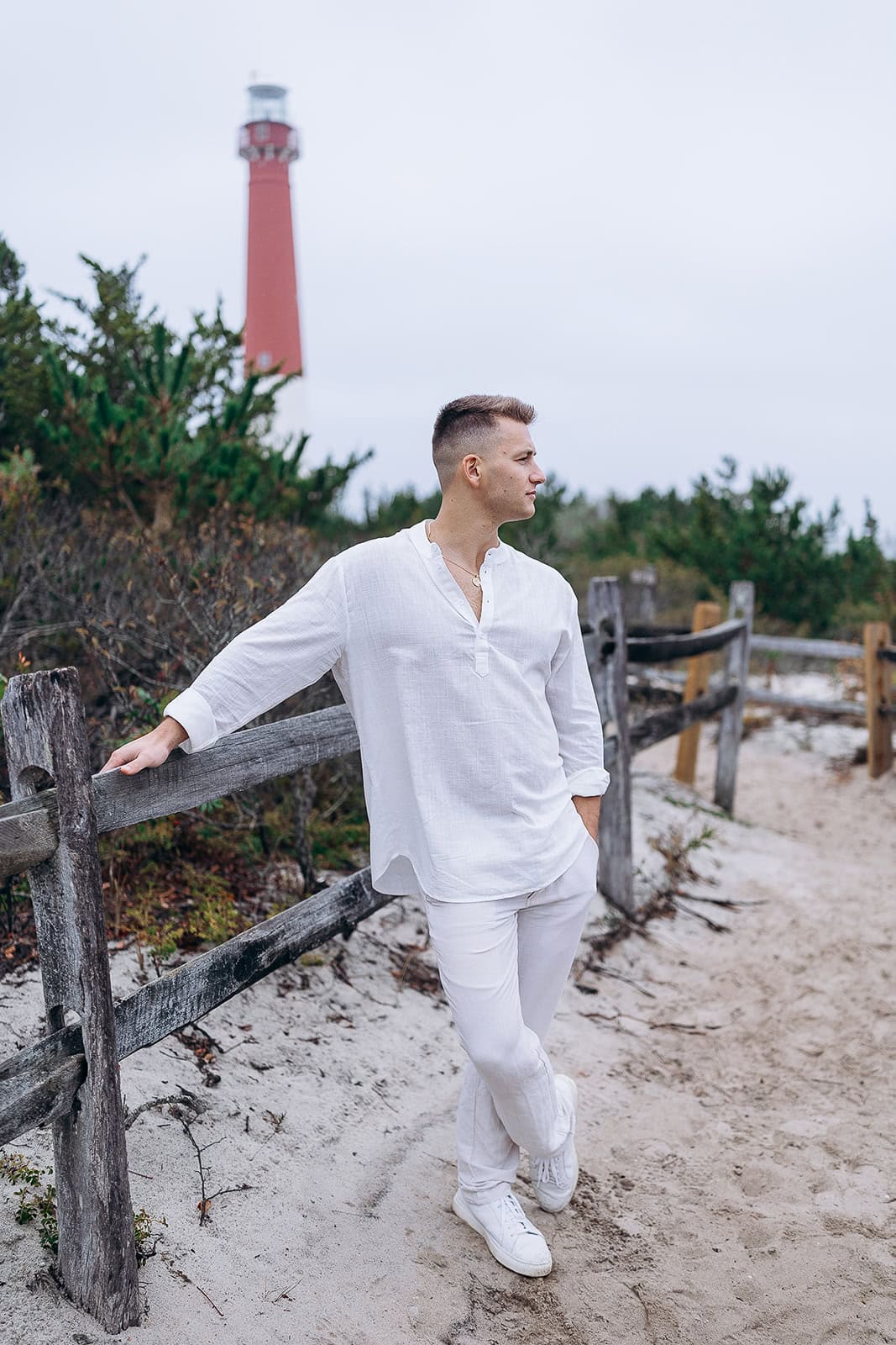 Man in white linen shirt and pants leaning on weathered wooden fence with Barnegat Light lighthouse in the background on a cloudy day.