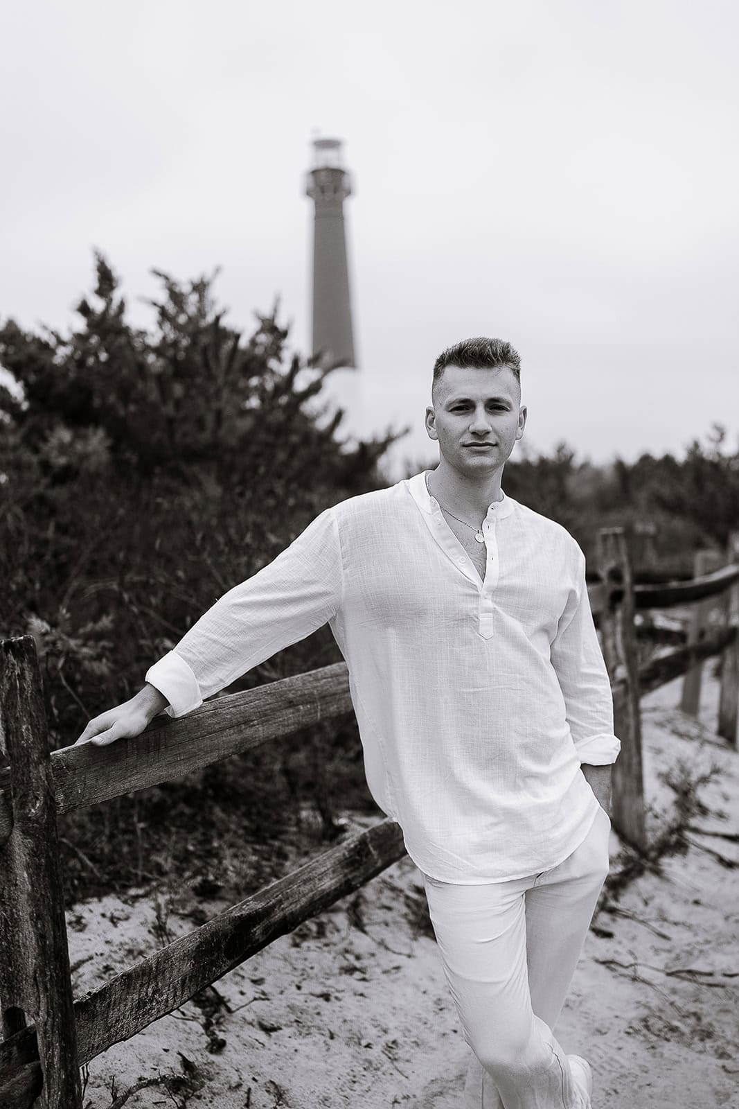Man leaning on wooden fence in white linen outfit with Barnegat Light lighthouse in the background on a sandy path.
