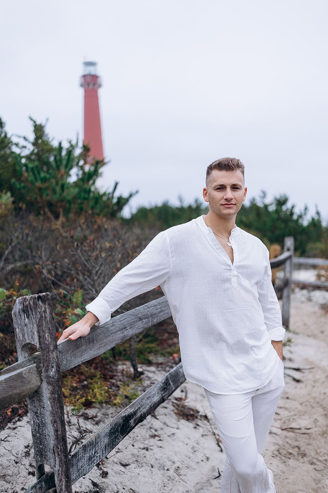 Man in white linen shirt and pants leaning on a rustic wooden fence on sandy ground with Barnegat Light lighthouse in the background under overcast sky.