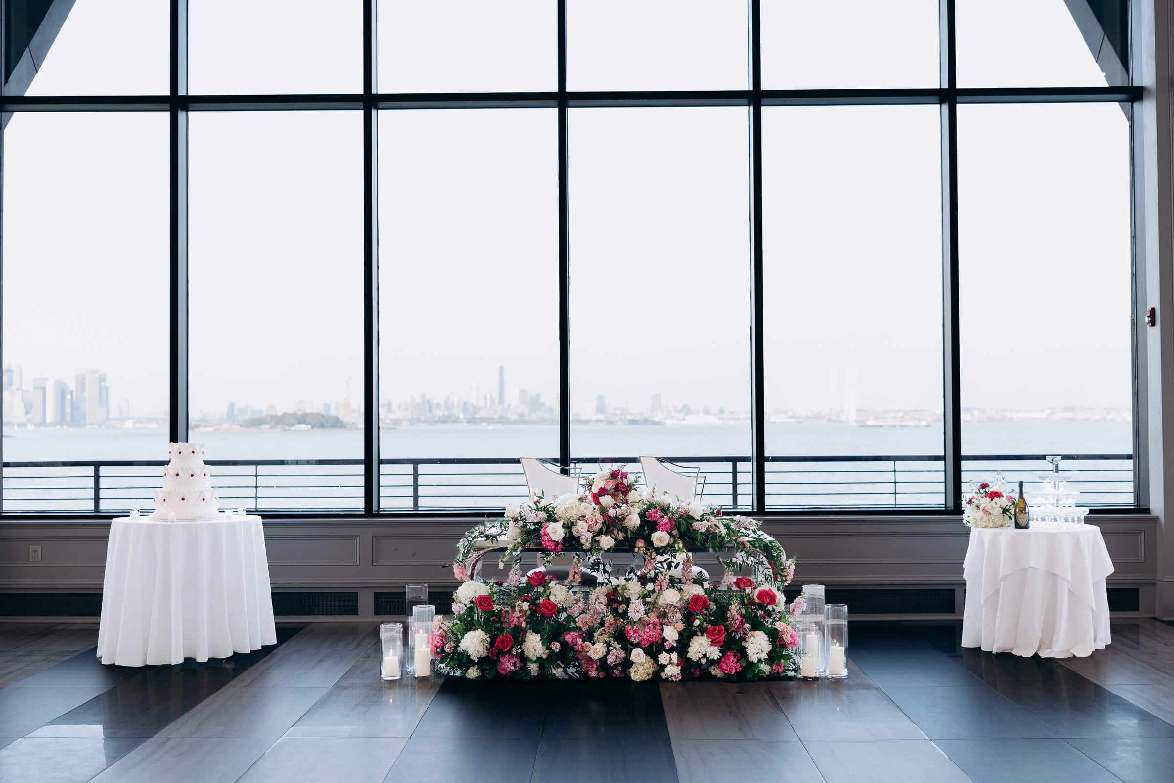 Sweetheart table with abundant pink and white flowers, tiered wedding cake on left, champagne tower on right, large windows with water view at Hudson House, Jersey City
