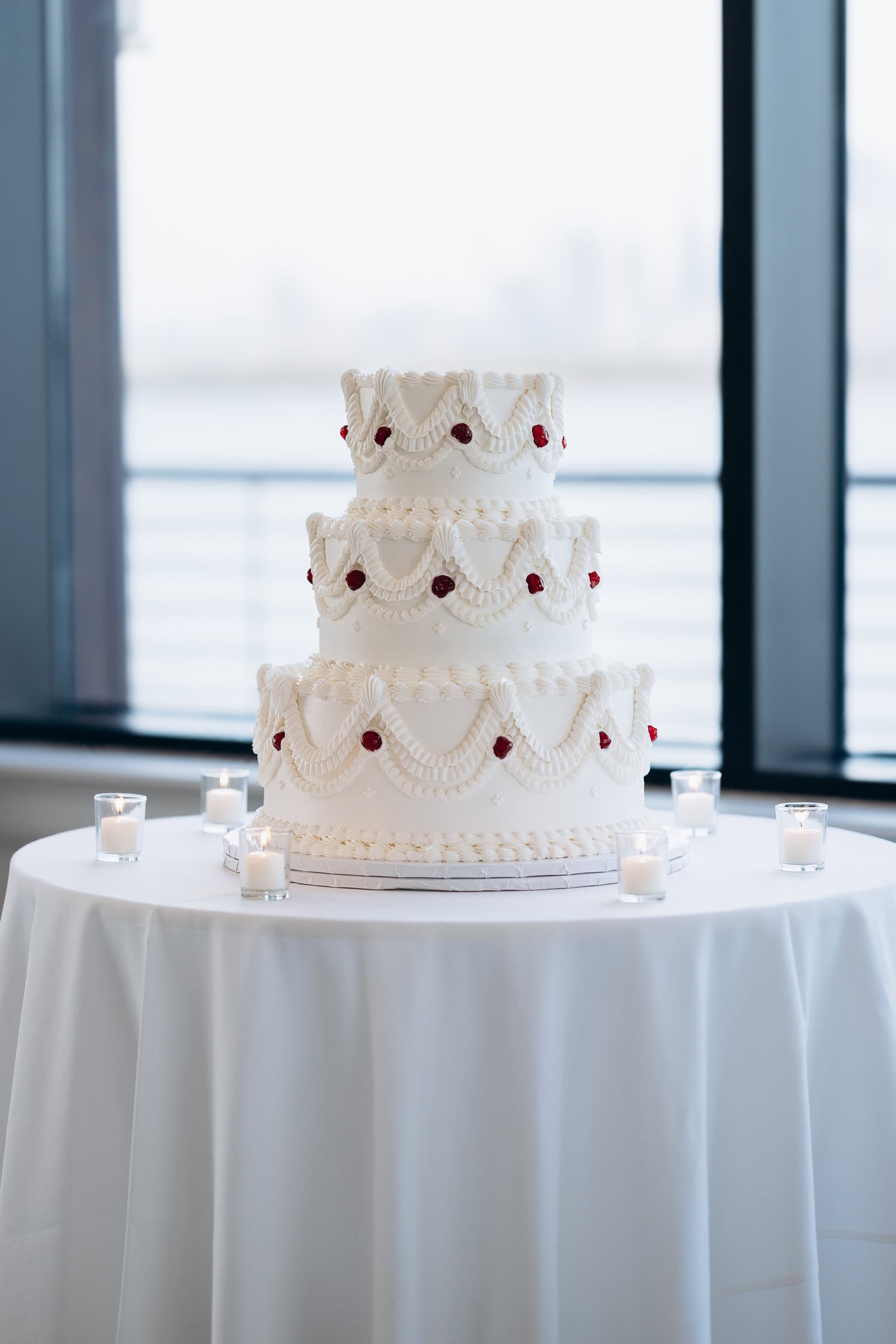 Three-tier white wedding cake with detailed piped swags and red dots on a white tablecloth, surrounded by small lit candles near large windows overlooking water at Hudson House, Jersey City.