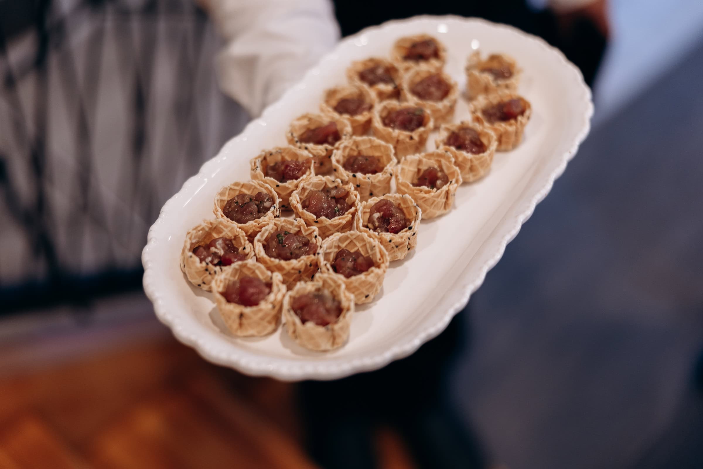 Close-up of a white platter holding multiple waffle cups filled with finely diced tuna tartare, held by a server at an event.