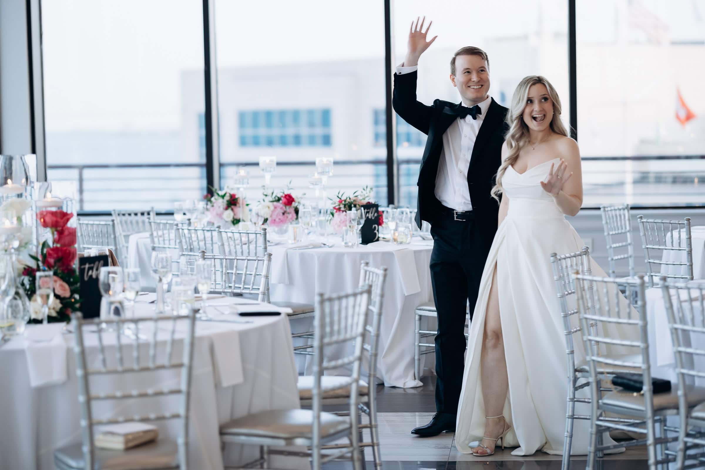 Bride and groom waving during their entrance at Hudson House, Jersey City, with elegantly set tables and waterfront view.